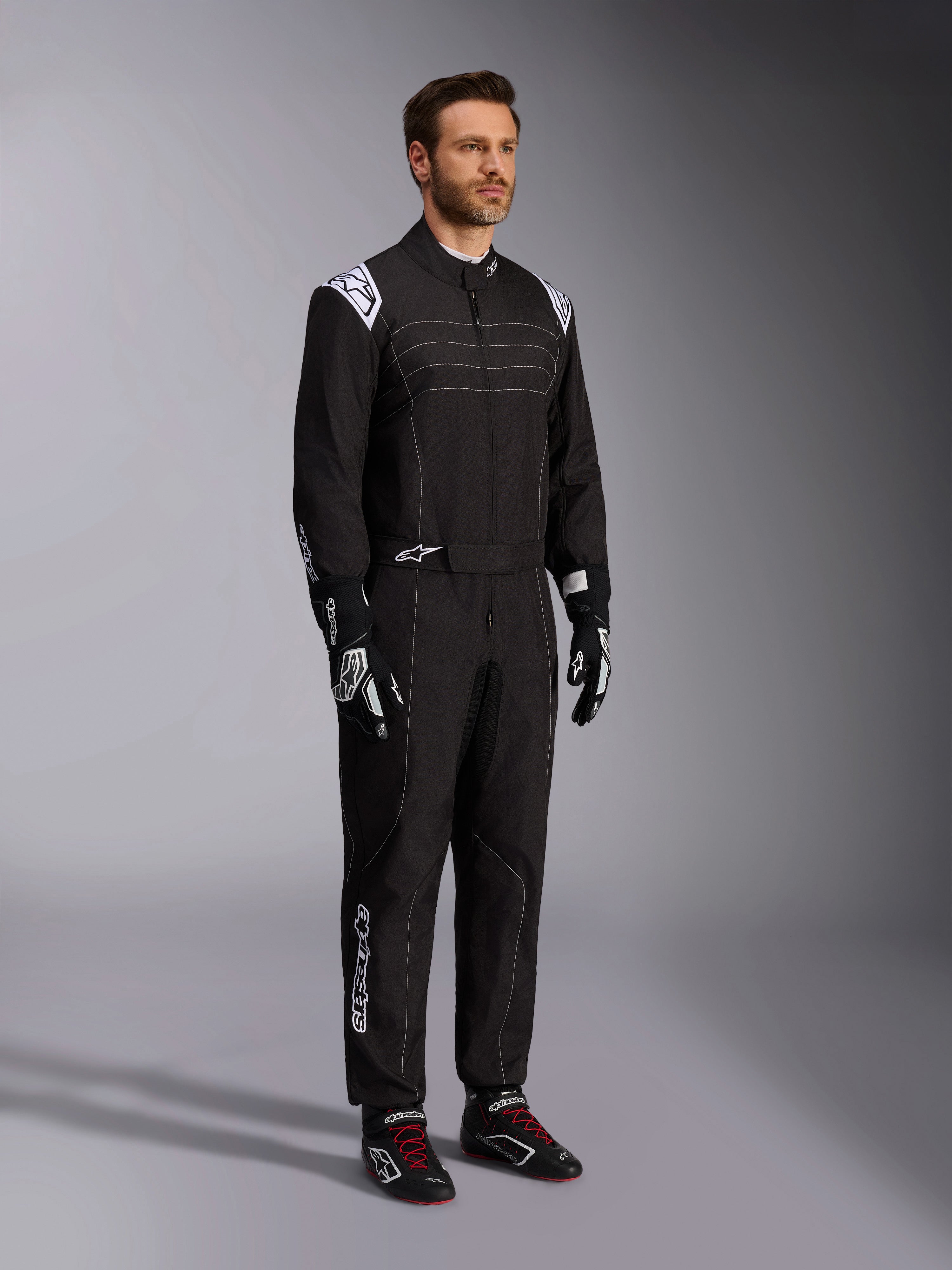 Alpinestars KMX-9 V3 Karting Suit, negro y blanco, modelo masculino vistiendo un mono de carreras completo con el logotipo de Alpinestars blanco en las hombreras, combinado con guantes de carreras negros y blancos y zapatillas de karting negras con cordones rojos, toma de estudio que muestra el perfil frontal y lateral