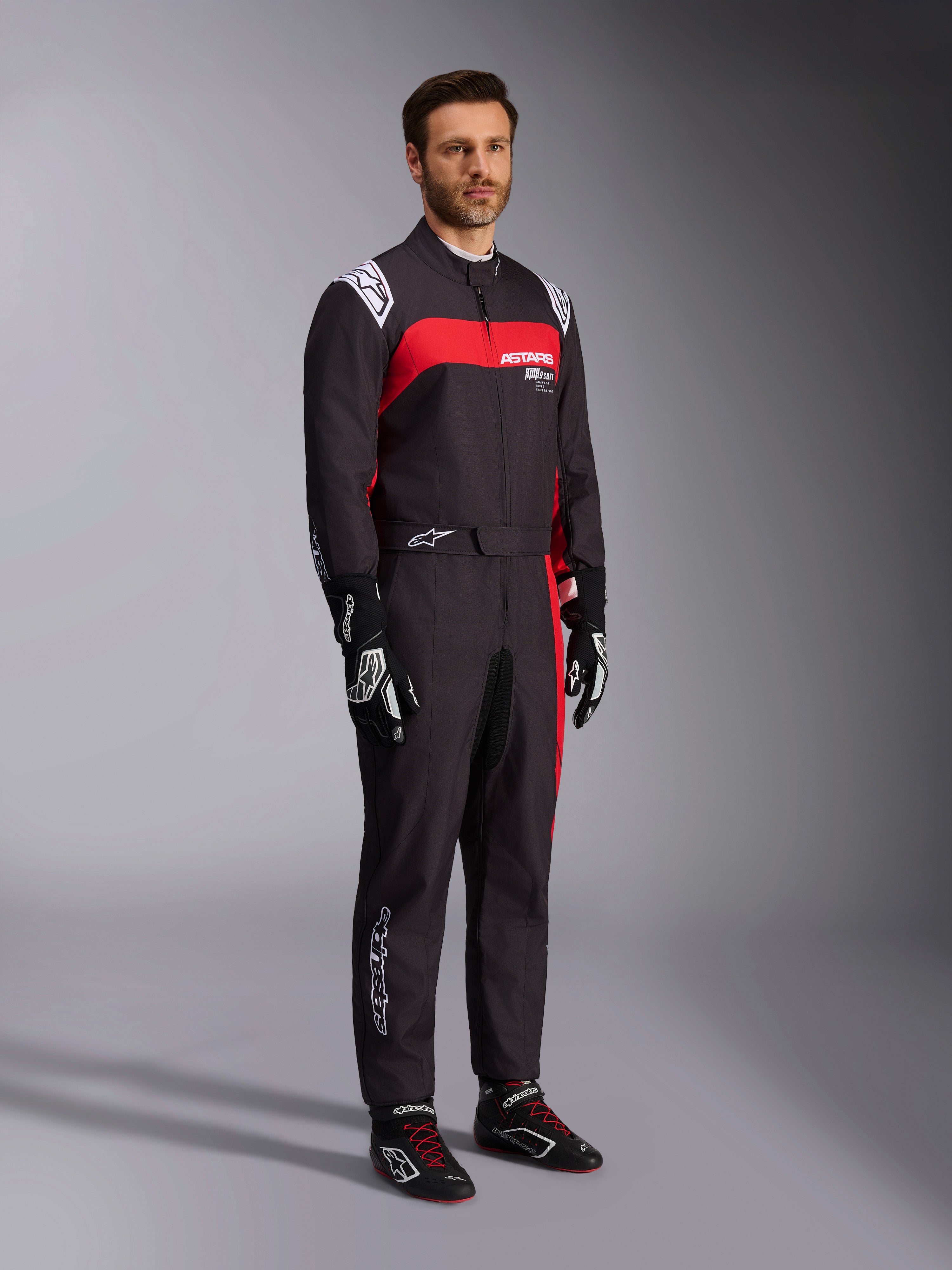 Modelo masculino con el mono Alpinestars KMX-9 V3 Graphic 3, traje de karting negro y rojo, con hombreras blancas y logotipo rojo en el pecho, combinado con guantes de carreras negros y calzado con cordones rojos, ropa profesional de automovilismo.