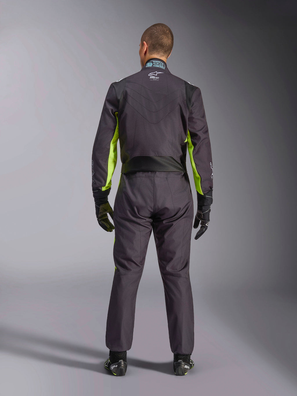 Vista posterior de cuerpo completo de una persona con el Traje Alpinestars KMX-9 V3 Graphic 3, mono de karting en color negro y amarillo flúor, que muestra los paneles elásticos ergonómicos en la espalda y los hombros, combinado con guantes de carreras negros y calzado.