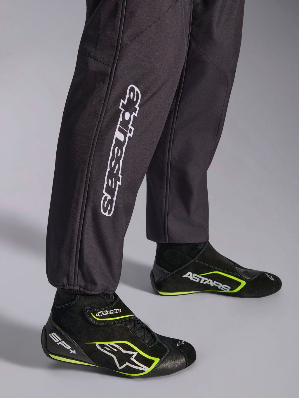 Mono Alpinestars KMX-9 V3 Graphic 3, mono de karting negro, primer plano de la parte inferior de las piernas y los puños de los tobillos, con marca Alpinestars vertical en blanco, combinado con calzado de karting en negro y amarillo flúor, detalle de equipamiento profesional de karting