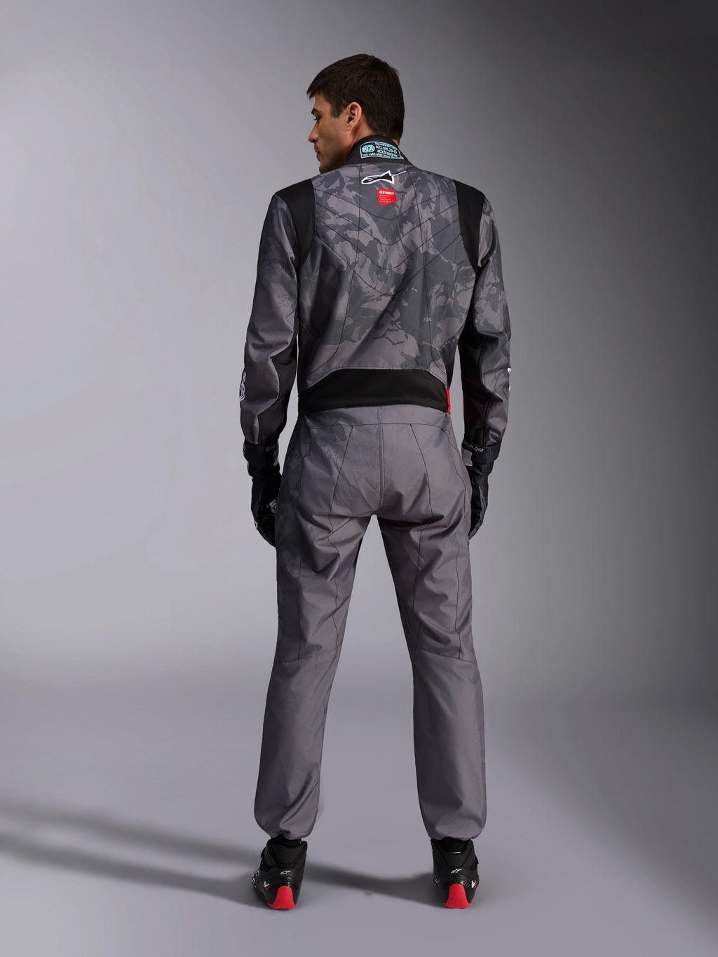 Alpinestars KMX-9 V3 Graphic 5 Suit, mono de karting, patrón de camuflaje en gris oscuro y negro en la parte superior, vista posterior que muestra los paneles elásticos y la marca Alpinestars, usado con guantes negros y calzado de carreras negro con detalles rojos, equipamiento profesional para automovilismo