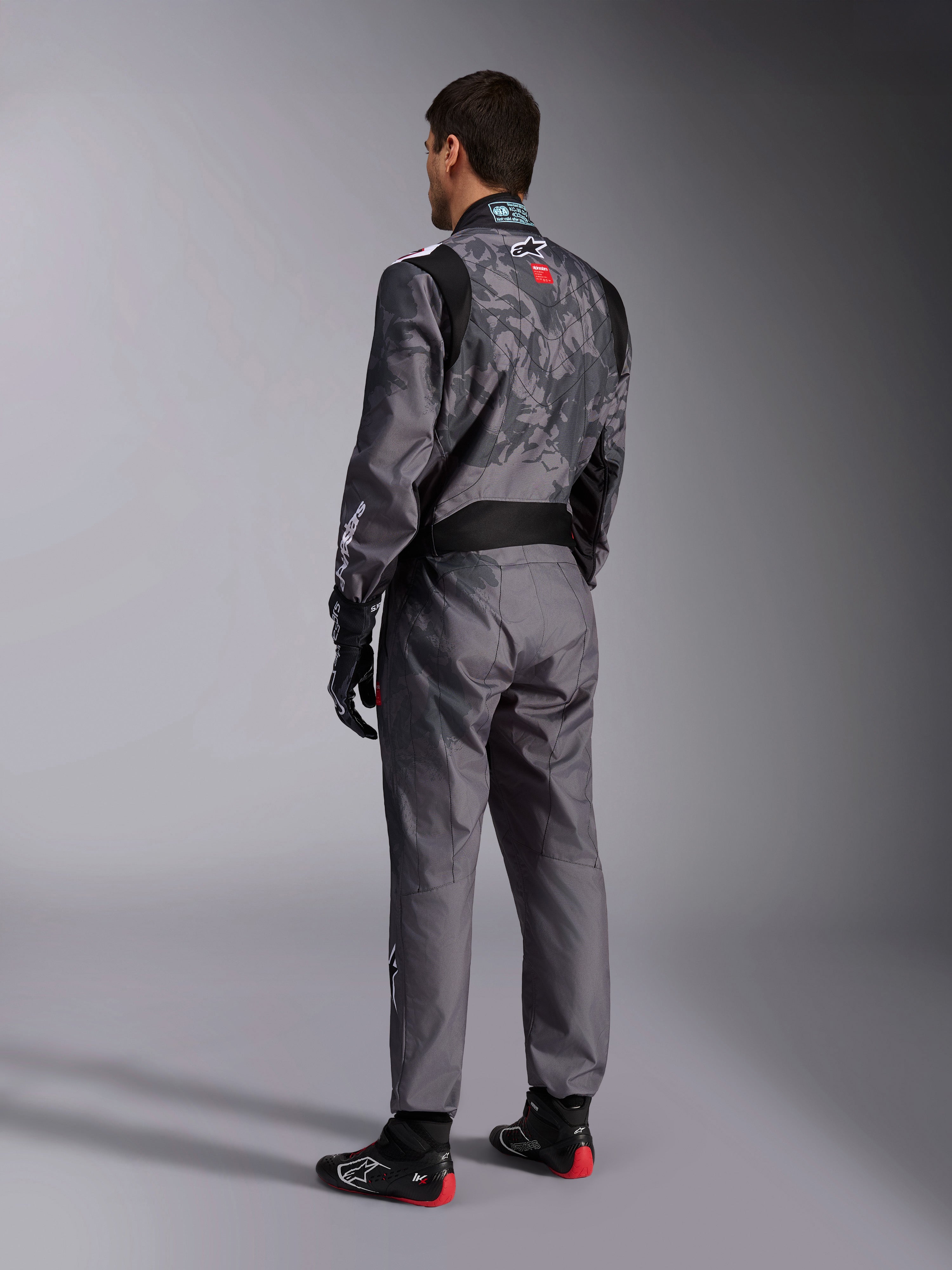 Alpinestars KMX-9 V3 Graphic 5 Suit, mono de karting en gris oscuro y negro, vista trasera de un modelo con el equipamiento de carreras completo que incluye guantes negros y rojos y calzado de karting, con patrones gráficos integrados y logotipos en la espalda.