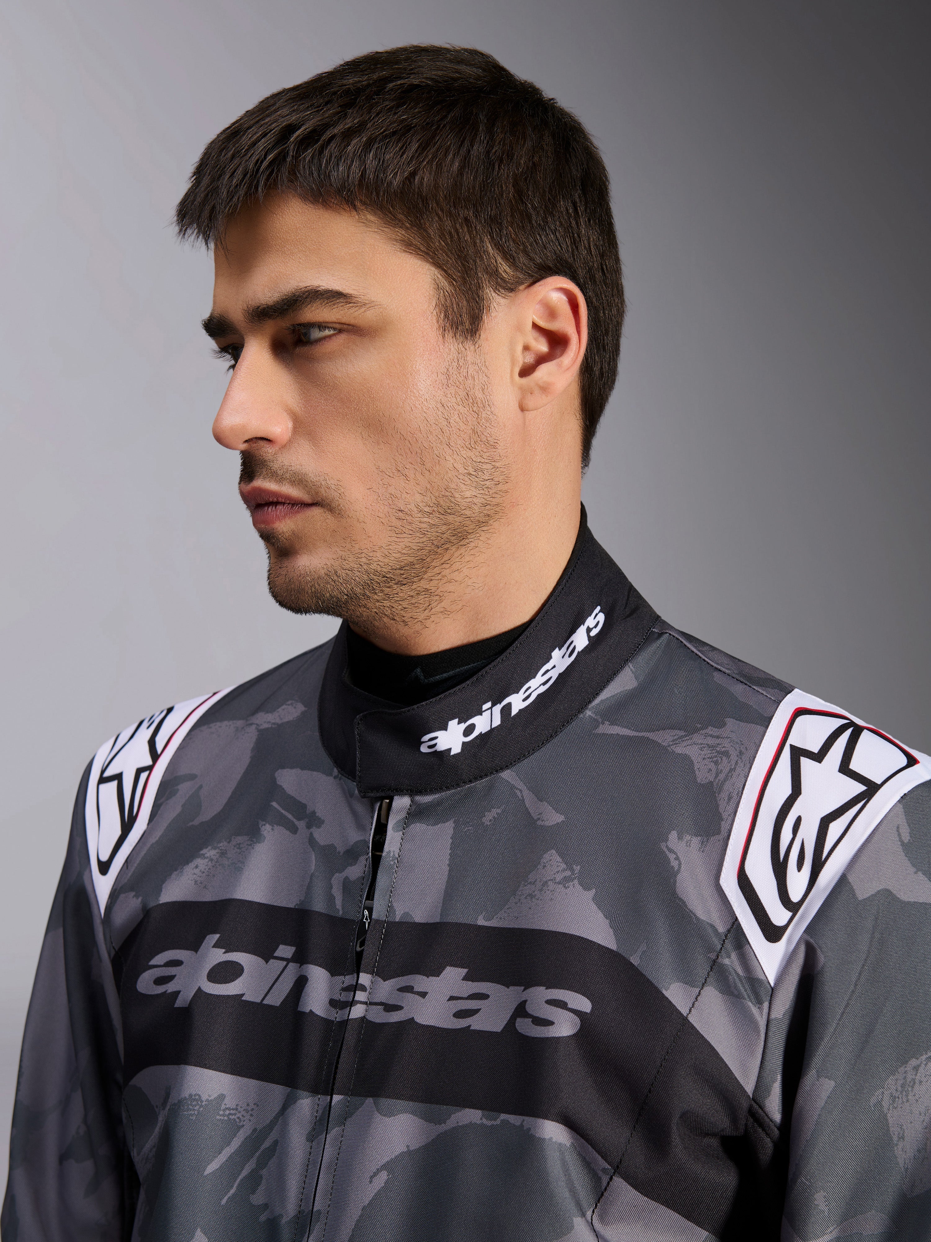 Alpinestars KMX-9 V3 Graphic 5 Suit, mono de karting, patrón de camuflaje gris oscuro y negro, primer plano del cuello y los hombros con logotipos blancos, indumentaria de carreras profesional