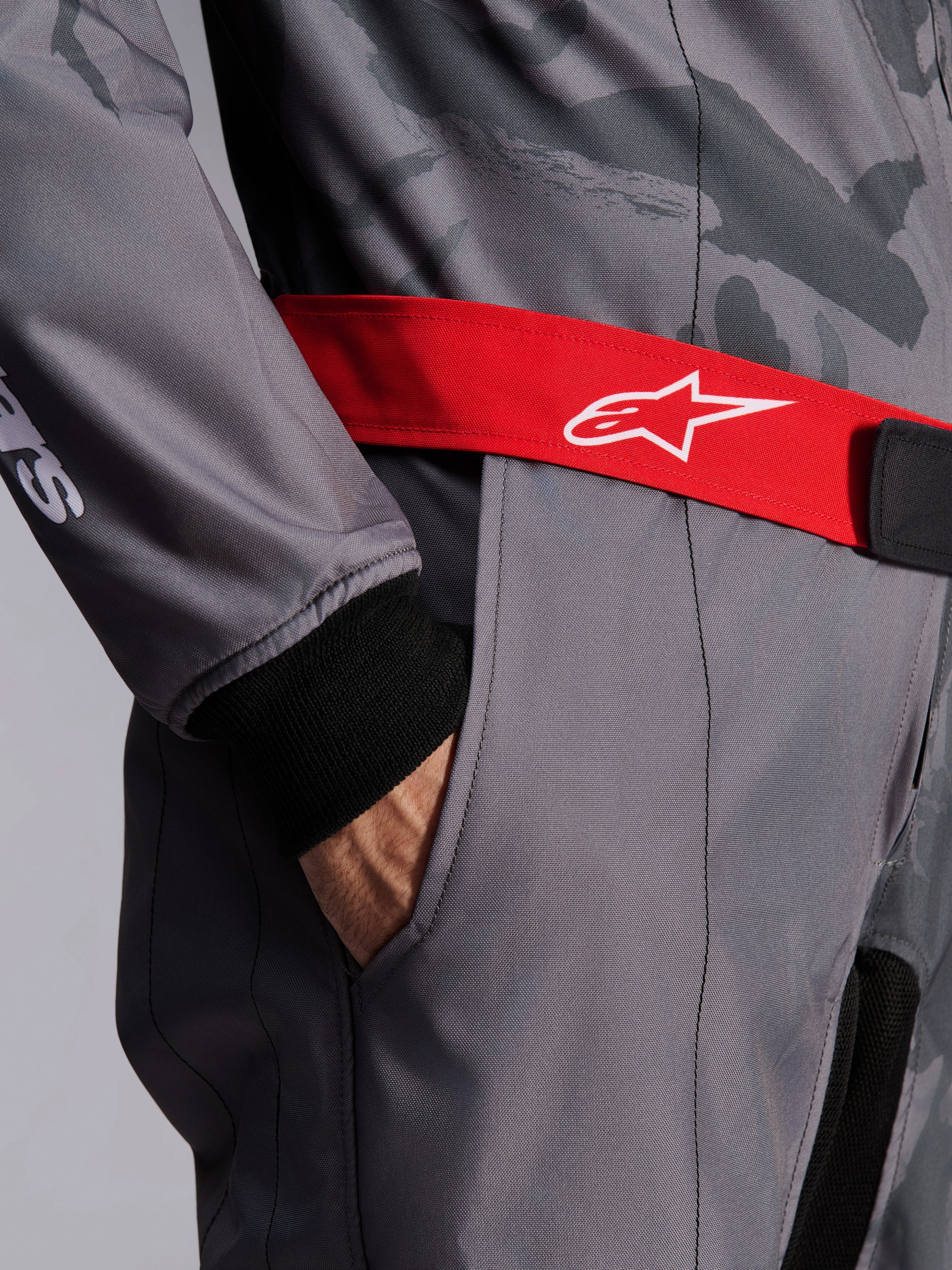 Alpinestars KMX-9 V3 Graphic 5 Suit, mono de karting, gris oscuro y negro con cinturón rojo, primer plano del cinturón ajustable rojo con logo blanco, mano en el bolsillo del muslo y puño de punto negro sobre tela con diseño gráfico gris