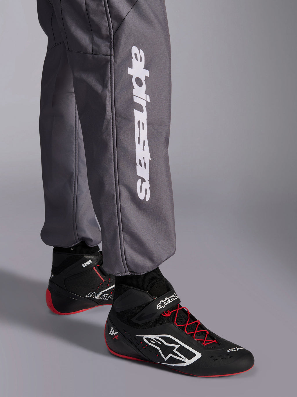 Mono Alpinestars KMX-9 V3 Graphic 5, mono de karting, gris oscuro, primer plano de la parte inferior de la pierna con el logotipo vertical de Alpinestars en blanco, combinado con calzado de carreras negro y rojo, material resistente para competición de karting