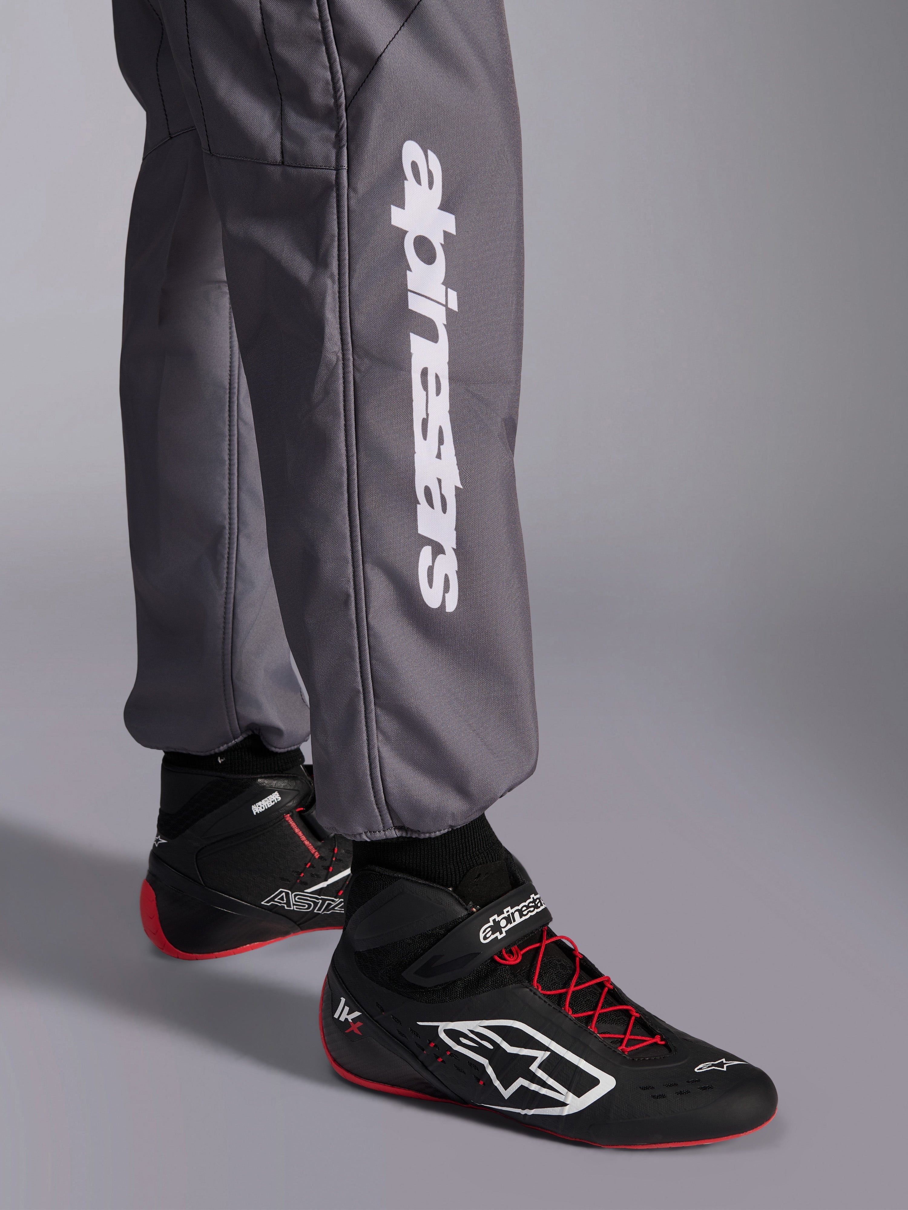 Mono Alpinestars KMX-9 V3 Graphic 5, mono de karting, gris oscuro, primer plano de la parte inferior de la pierna con el logotipo vertical de Alpinestars en blanco, combinado con calzado de carreras negro y rojo, material resistente para competición de karting