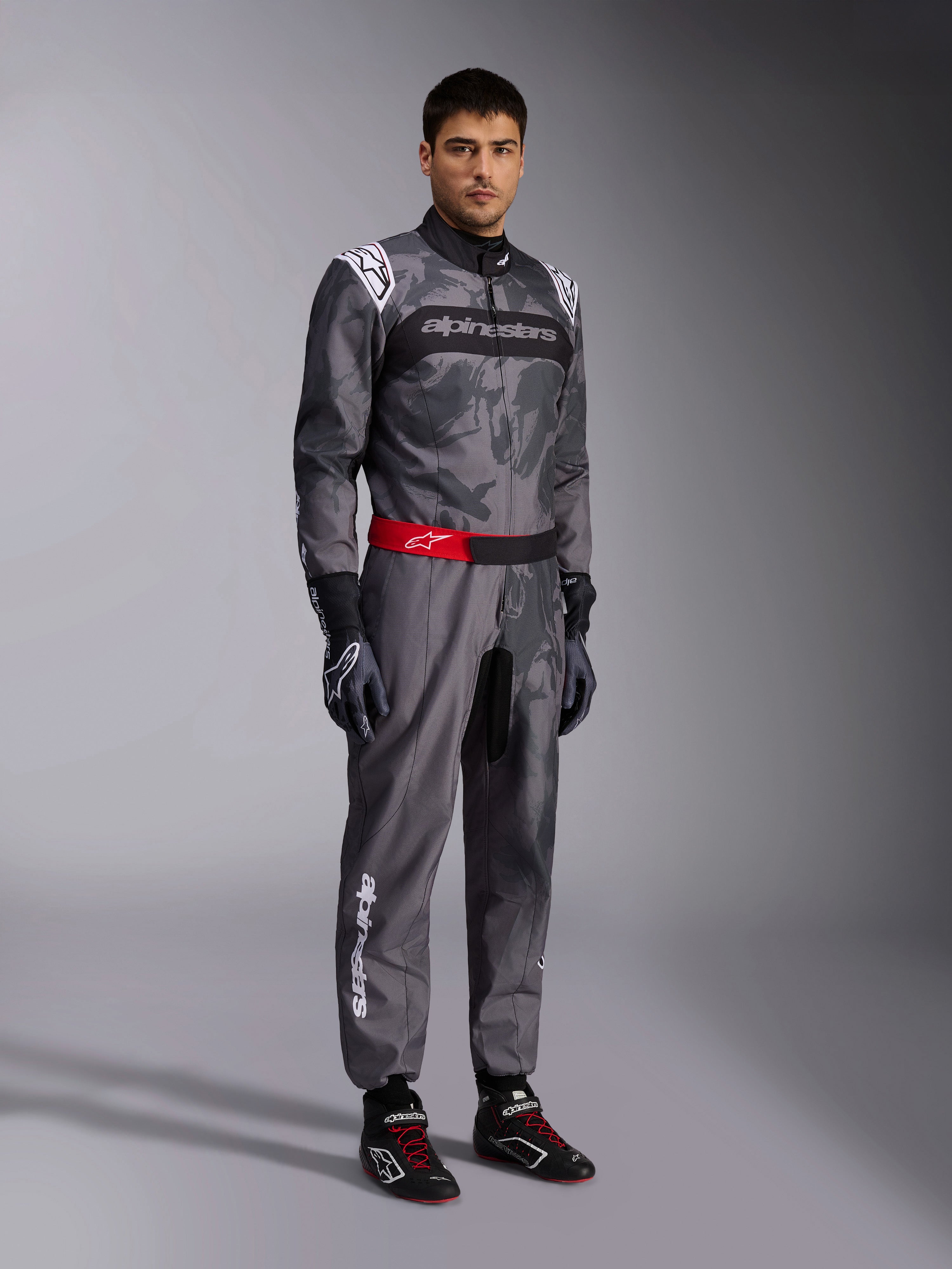 Mono Alpinestars KMX-9 V3 Graphic 5, traje de karting gris oscuro y negro con patrón de camuflaje, cinturón rojo con logo blanco, combinado con guantes y botas de carreras, equipamiento profesional de automovilismo para karting