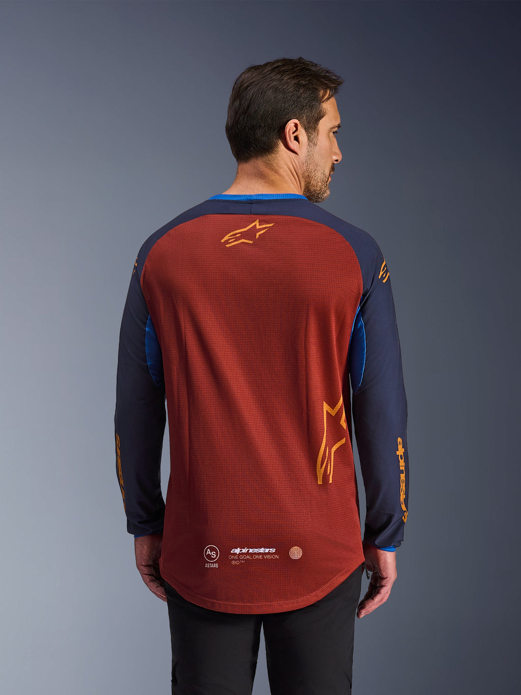 Alpinestars Maxdura Dual Jersey en Rust Brown y Mood Indigo, vista desde atrás, con torso de malla transpirable, mangas azul oscuro con logotipos de Alpinestars en naranja y dobladillo trasero caído para mayor cobertura.