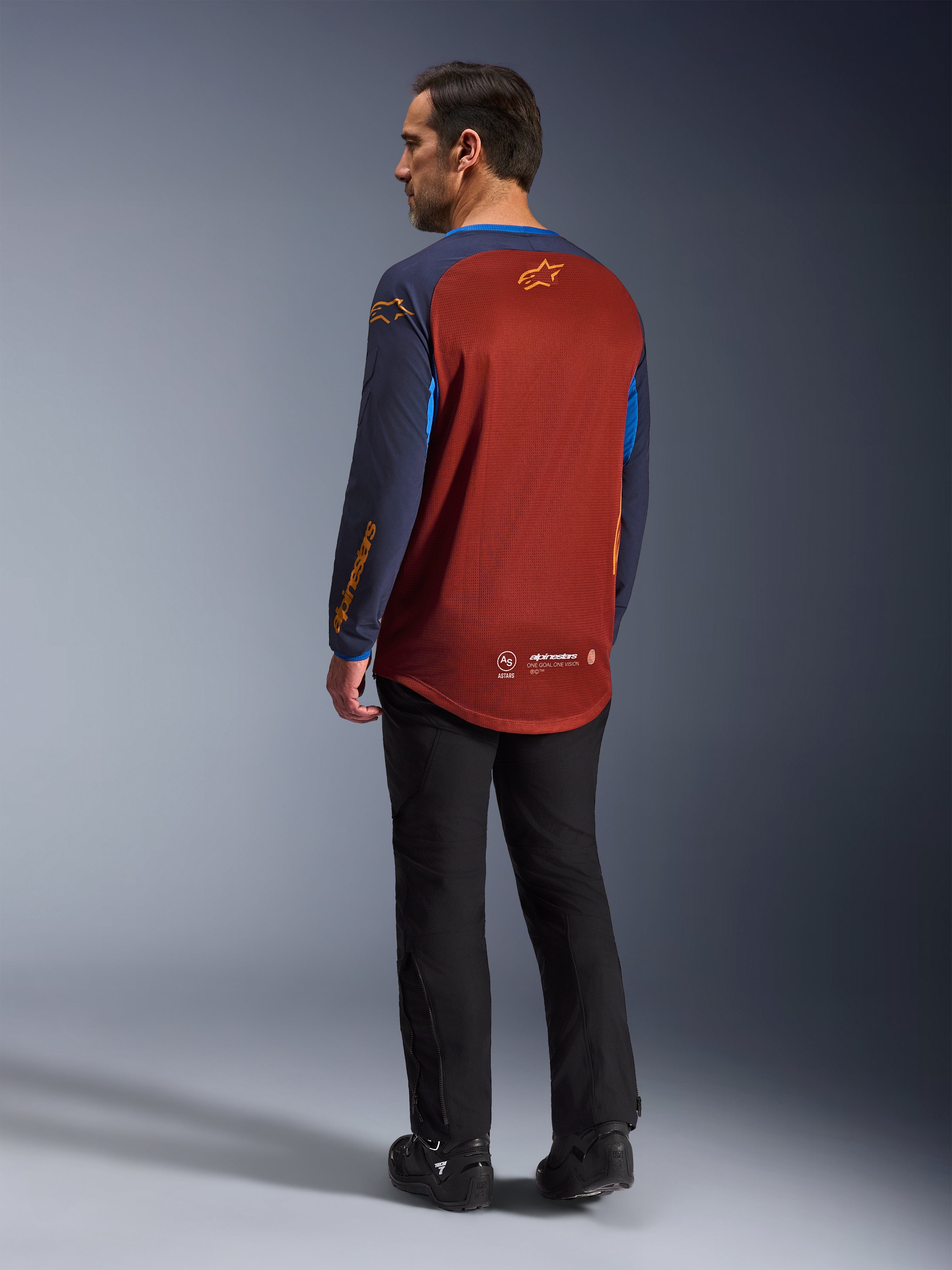 Alpinestars Maxdura Dual Jersey, Marrón Óxido y Azul Índigo, jersey de aventura para motociclismo visto desde atrás, panel de malla de color óxido para ventilación, mangas azul marino con logotipos naranjas, usado con pantalones y botas de montar negros, equipamiento técnico para off-road