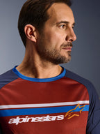 Alpinestars Maxdura Dual Jersey, marrón óxido y azul Mood Indigo, primer plano de un piloto usando el jersey de malla técnica, con franjas horizontales blancas y azules en el pecho, marca Alpinestars en blanco y logotipo de estrella naranja.