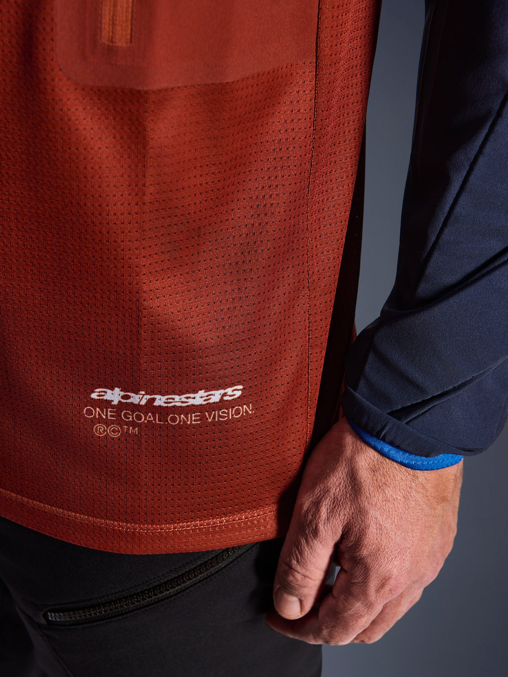 Alpinestars Maxdura Dual Jersey, jersey de motociclismo de aventura, marrón óxido y azul índigo, primer plano que muestra el tejido de malla del torso y la manga tejida azul marino, marca blanca Alpinestars 'One Goal One Vision' en el dobladillo inferior.