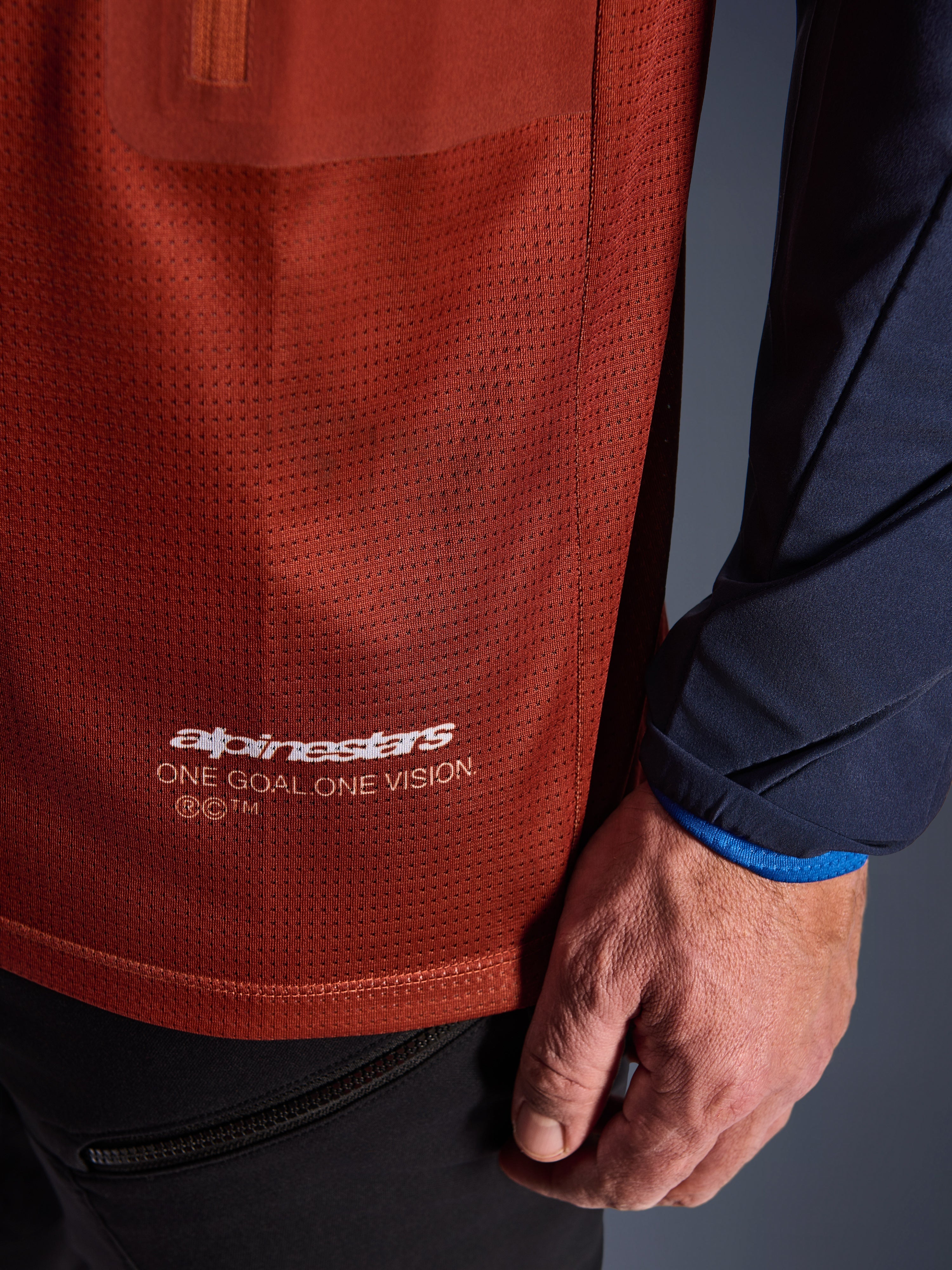 Alpinestars Maxdura Dual Jersey, jersey de motociclismo de aventura, marrón óxido y azul índigo, primer plano que muestra el tejido de malla del torso y la manga tejida azul marino, marca blanca Alpinestars 'One Goal One Vision' en el dobladillo inferior.