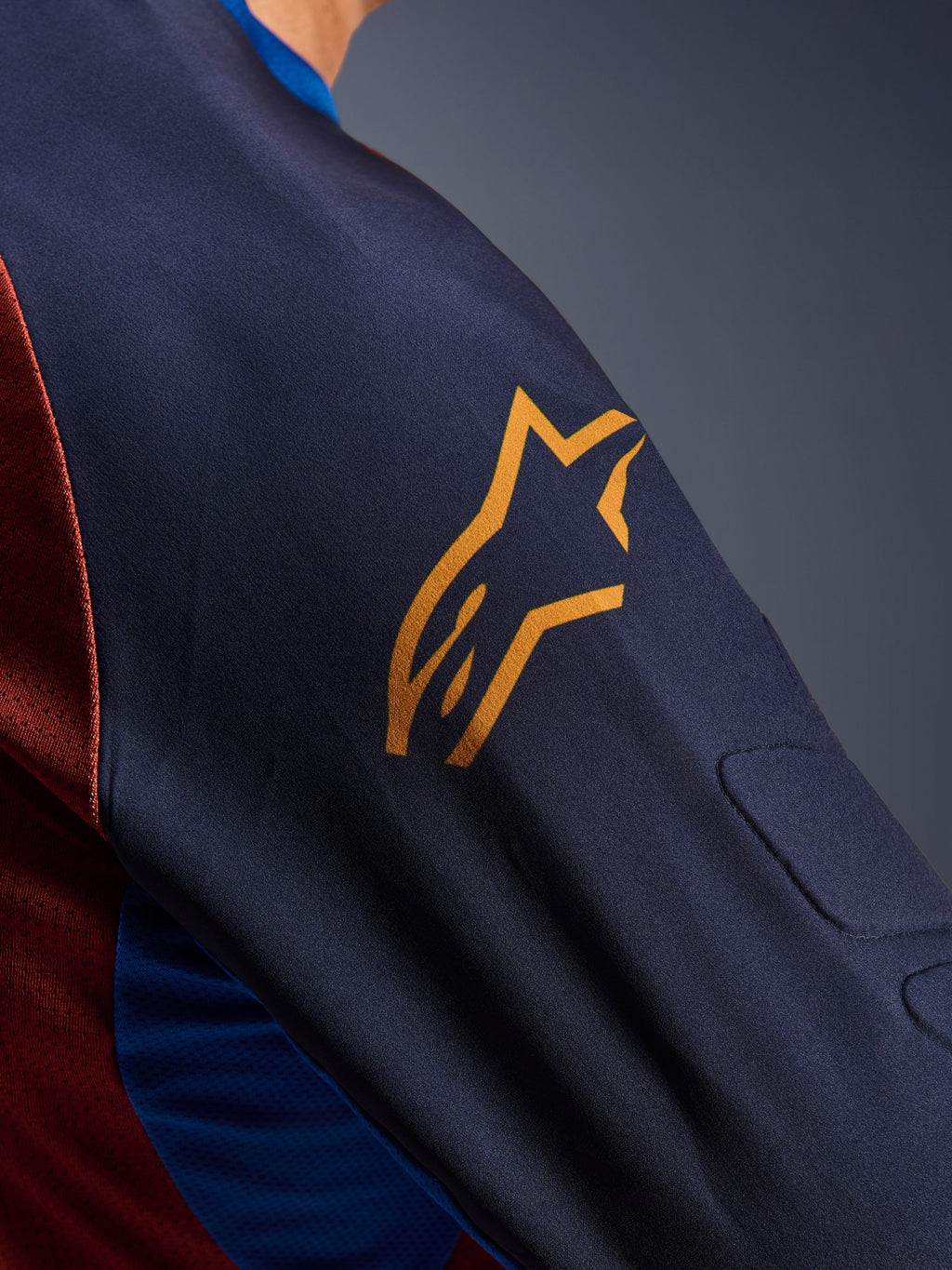 Alpinestars Maxdura Dual Jersey, primer plano de la manga en color Mood Indigo con un logotipo amarillo de Alpinestars, que muestra el acolchado de malla espaciadora integrado y el tejido de malla del torso en color Rust Brown, prenda técnica de motociclismo de aventura