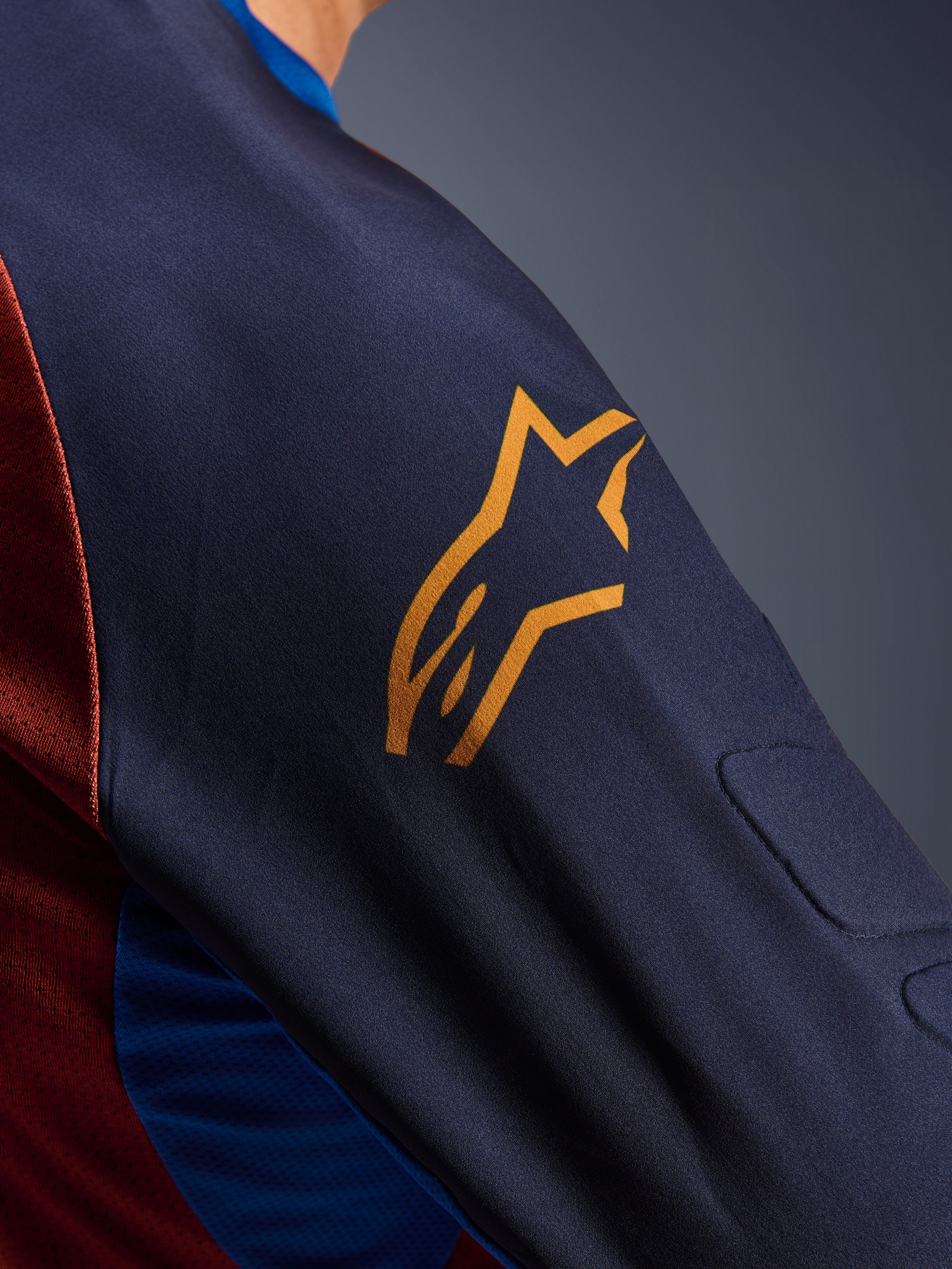 Alpinestars Maxdura Dual Jersey, primer plano de la manga en color Mood Indigo con un logotipo amarillo de Alpinestars, que muestra el acolchado de malla espaciadora integrado y el tejido de malla del torso en color Rust Brown, prenda técnica de motociclismo de aventura