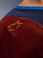 Primer plano del Alpinestars Maxdura Dual Jersey, tejido de malla marrón óxido en la parte trasera, pequeño logotipo de estrella naranja, panel superior azul oscuro y ribete del cuello en azul brillante, vista detallada de jersey de moto de aventura