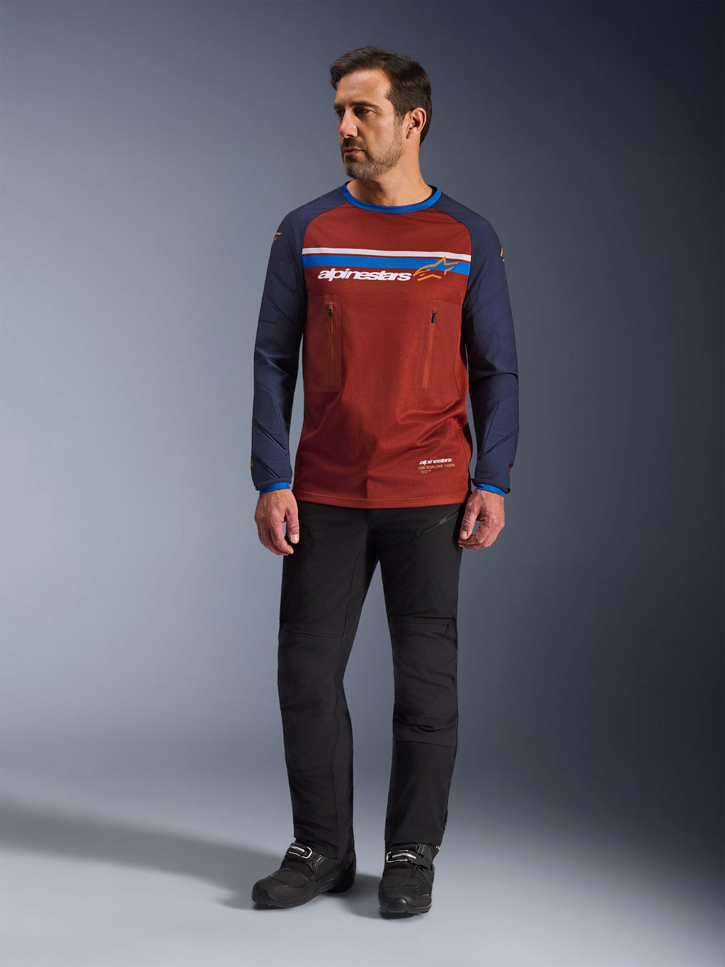 Alpinestars Maxdura Dual Jersey, marrón óxido y azul Mood Indigo, jersey de motociclismo de aventura con torso de malla y mangas azul marino, usado por un modelo con pantalones de motociclismo negros y botas negras, equipamiento técnico dual-sport para conducción fuera de carretera