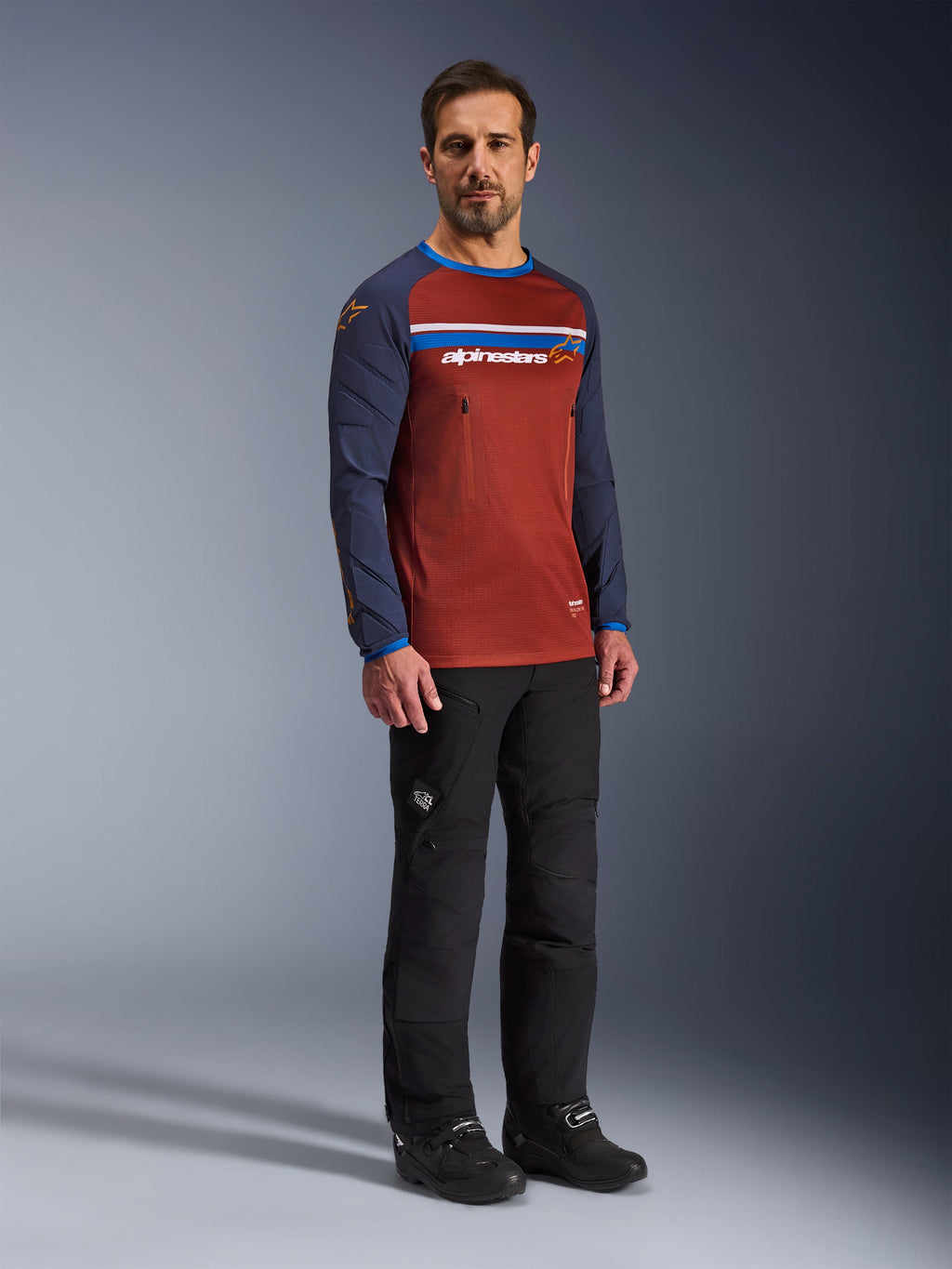 Hombre con el Jersey Alpinestars Maxdura Dual en Rust Brown y Mood Indigo, con torso de malla y mangas acolchadas, combinado con pantalones de conducción y botas negros, equipamiento de motociclismo de aventura.