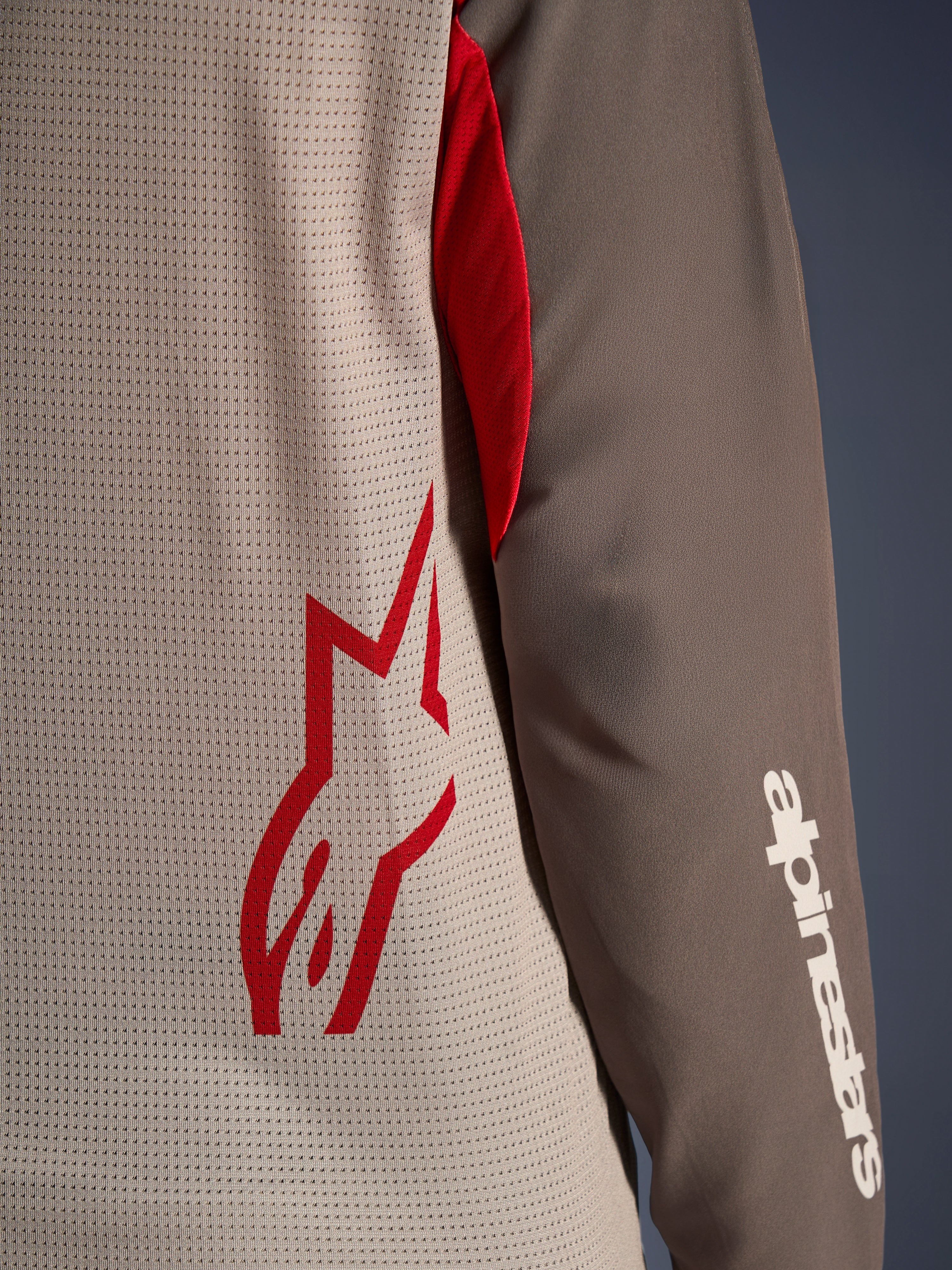 Alpinestars Maxdura Dual Jersey, Aluminum Walnut, vista de cerca que muestra el torso de malla ventilada con un logo de estrella roja y una manga marrón resistente a la abrasión con la marca Alpinestars en blanco, equipamiento técnico de moto para aventura y dual-sport