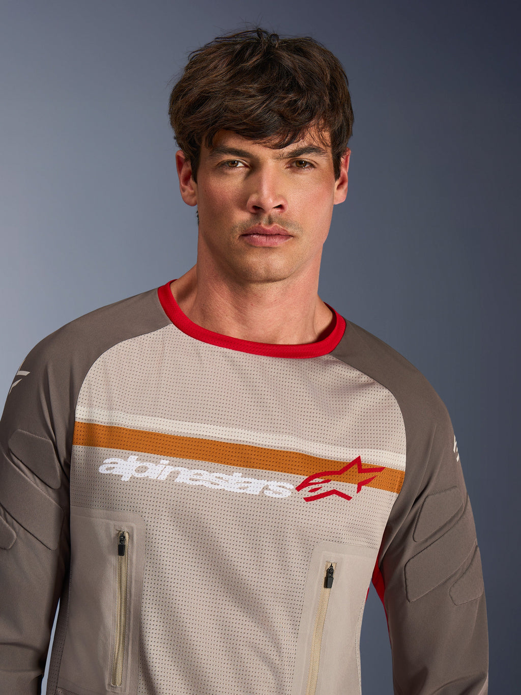 Modelo masculino vistiendo el jersey Alpinestars Maxdura Dual en Aluminum Walnut, jersey de motocicleta de aventura con torso de malla completa, mangas resistentes a la abrasión, cuello rojo y bolsillos con cremallera en el pecho, con franjas en el pecho de color naranja y blanco y el logotipo de Alpinestars.