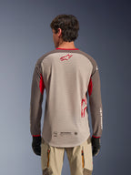 Vista trasera de un hombre con el jersey Alpinestars Maxdura Dual en color Aluminum Walnut, jersey de moto de aventura con torso de malla color canela y logo de estrella rojo, mangas marrón oscuro con detalles rojos, combinado con pantalones beige y guantes negros, diseñado para ventilación off-road de alto rendimiento.