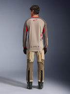 Alpinestars Maxdura Dual Jersey en Aluminum Walnut, vista trasera, torso de malla beige con mangas marrones y logotipos rojos, dobladillo trasero extendido, combinado con pantalones de conducción de aventura y botas negras.