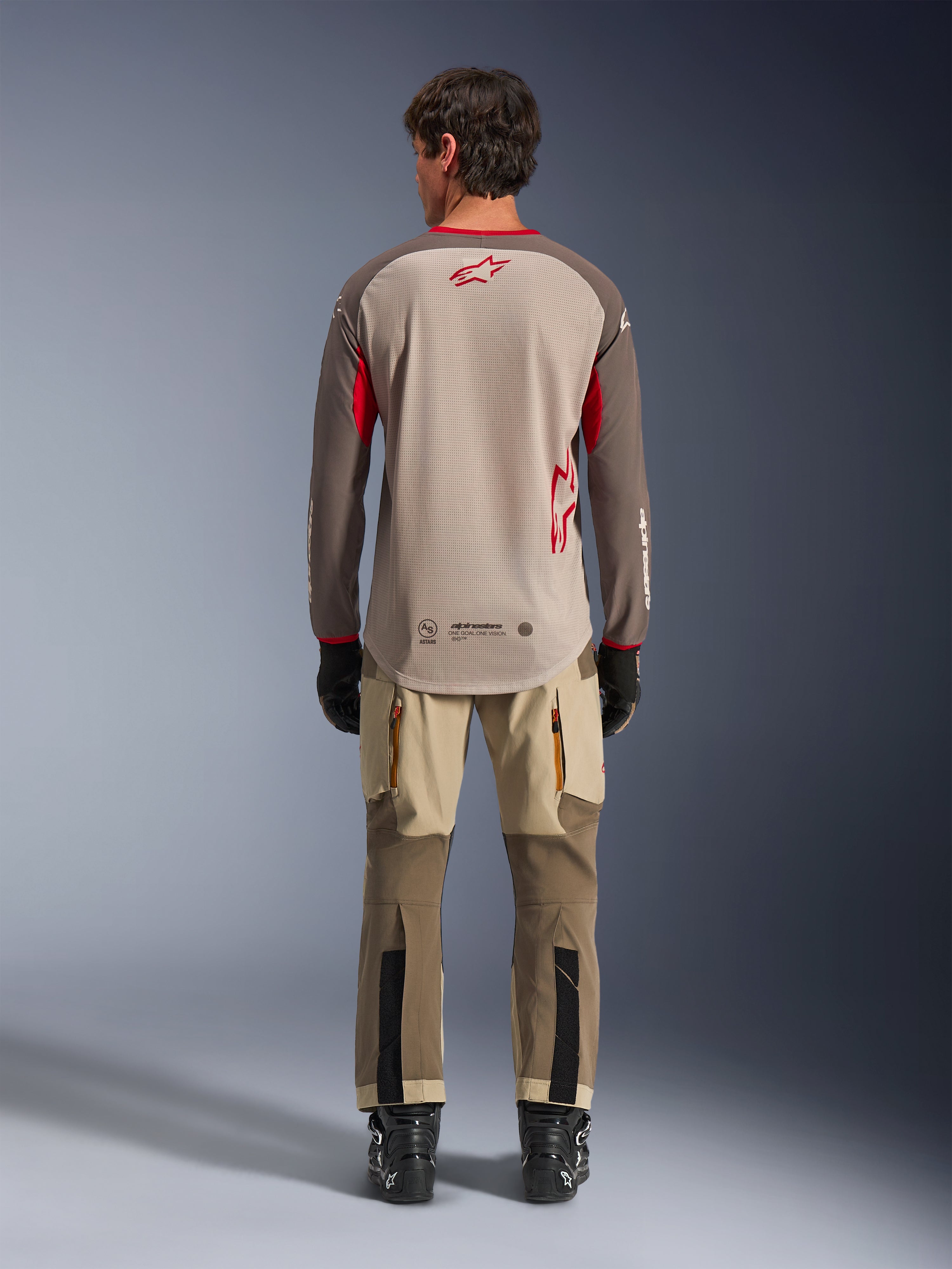 Alpinestars Maxdura Dual Jersey en Aluminum Walnut, vista trasera, torso de malla beige con mangas marrones y logotipos rojos, dobladillo trasero extendido, combinado con pantalones de conducción de aventura y botas negras.