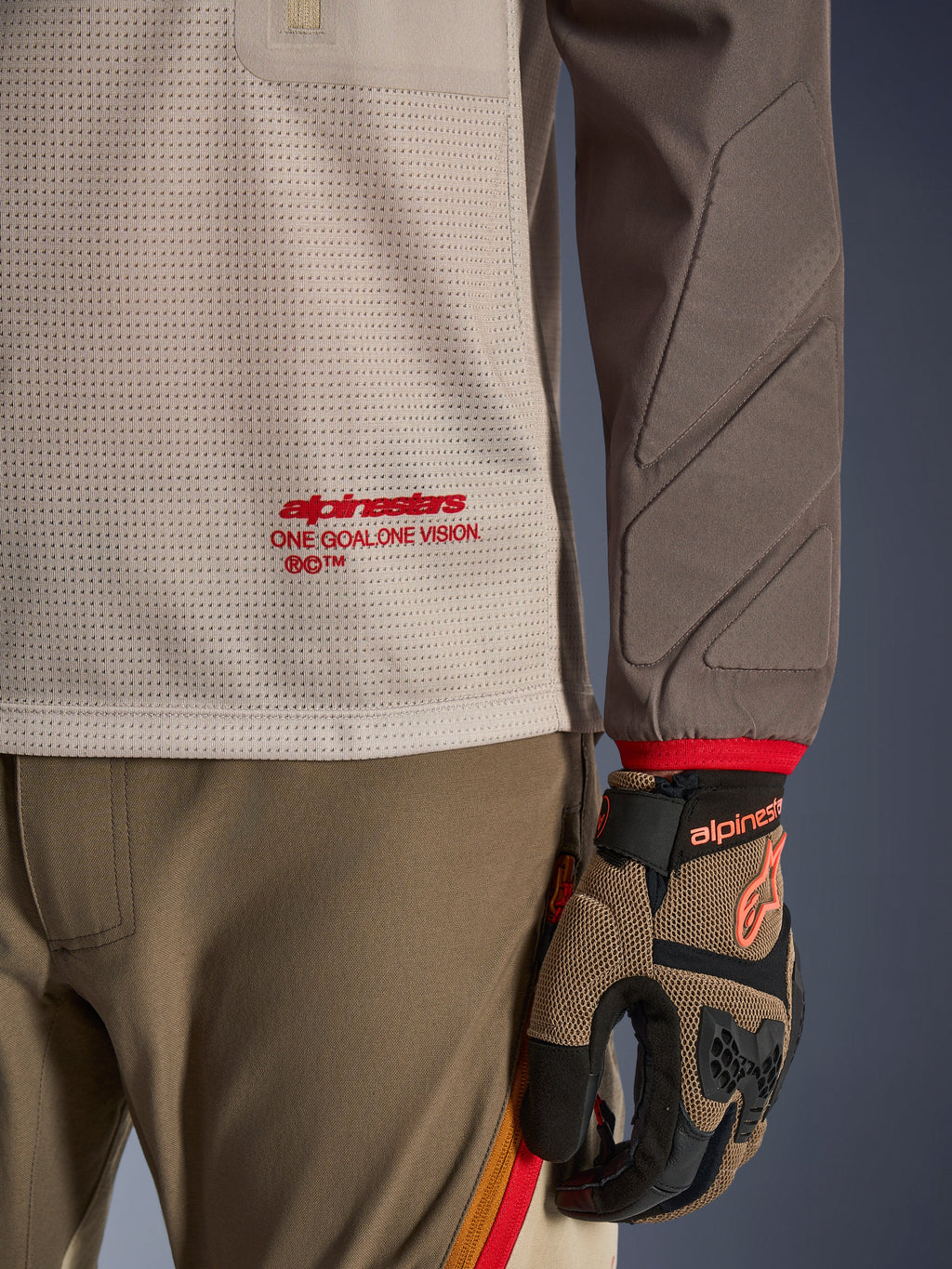 Alpinestars Maxdura Dual Jersey en Aluminum Walnut, vista detallada que muestra el torso de malla ventilada con logotipo en rojo, manga resistente a la abrasión con acolchado integrado, combinada con pantalones y guantes de conducción de aventura a juego