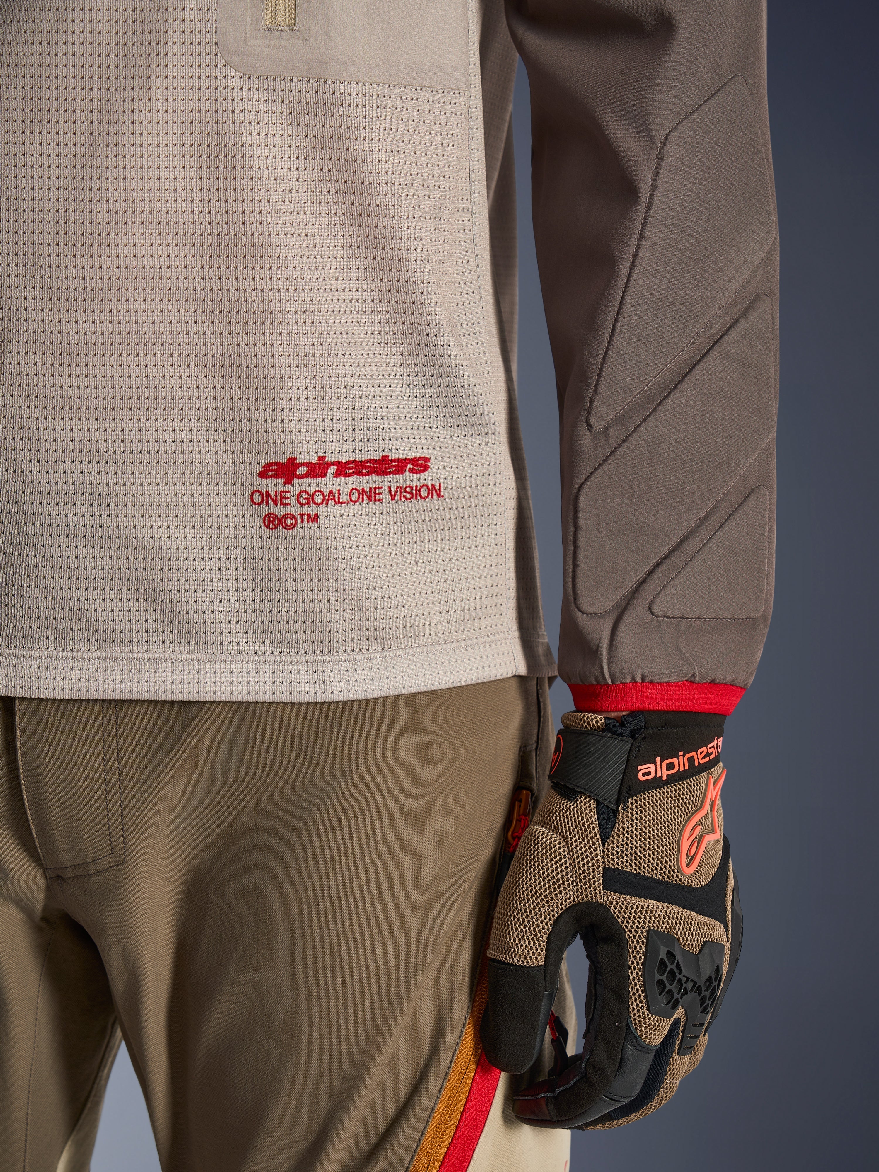 Alpinestars Maxdura Dual Jersey en Aluminum Walnut, vista detallada que muestra el torso de malla ventilada con logotipo en rojo, manga resistente a la abrasión con acolchado integrado, combinada con pantalones y guantes de conducción de aventura a juego