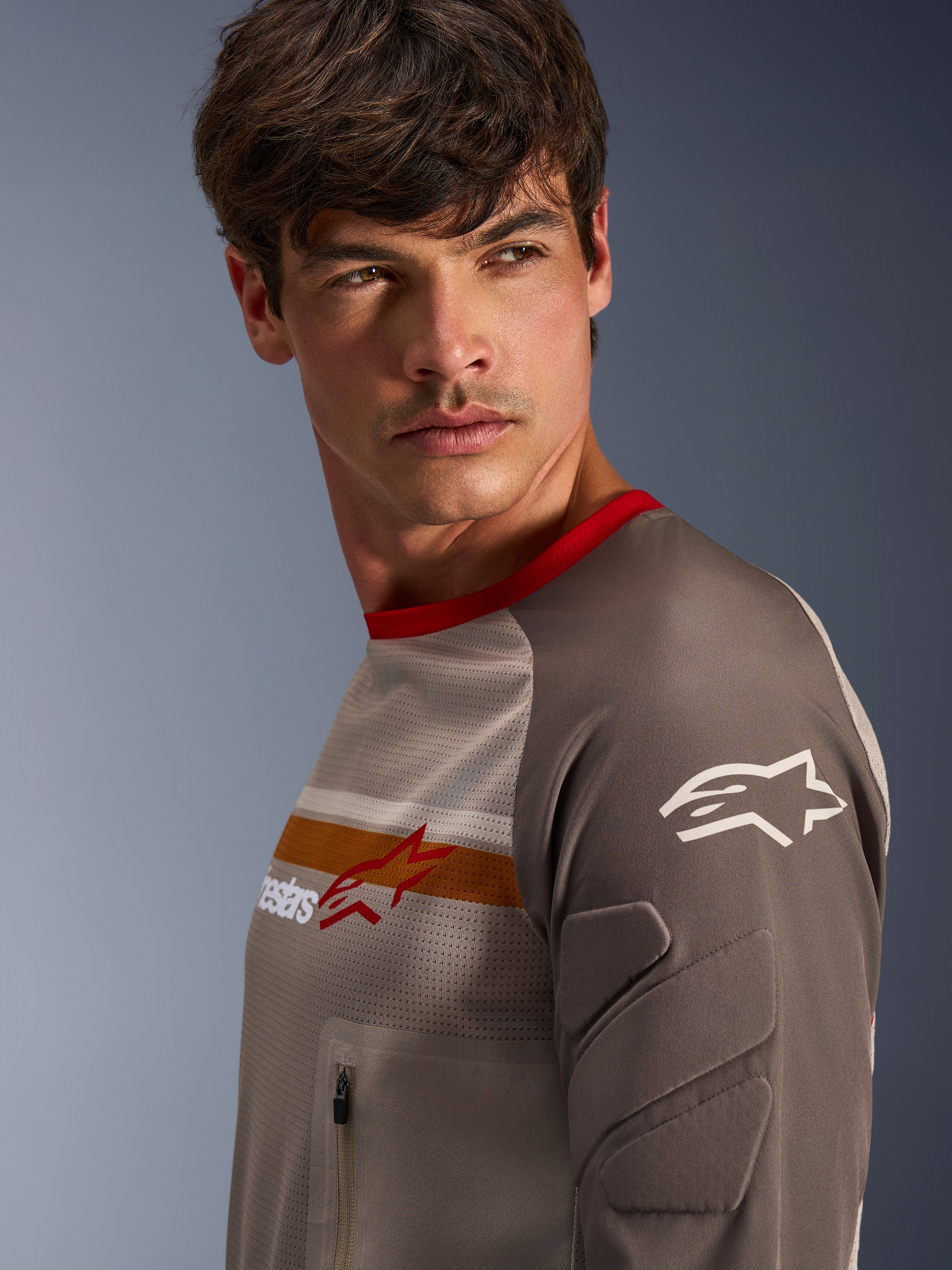 Modelo masculino vistiendo el jersey Alpinestars Maxdura Dual en color Aluminum Walnut, con torso de malla ventilada, cuello rojo y franja naranja en el pecho. Vista de primer plano de la manga resistente a la abrasión con acolchado integrado y bolsillo con cremallera en el pecho para conducción de aventura.