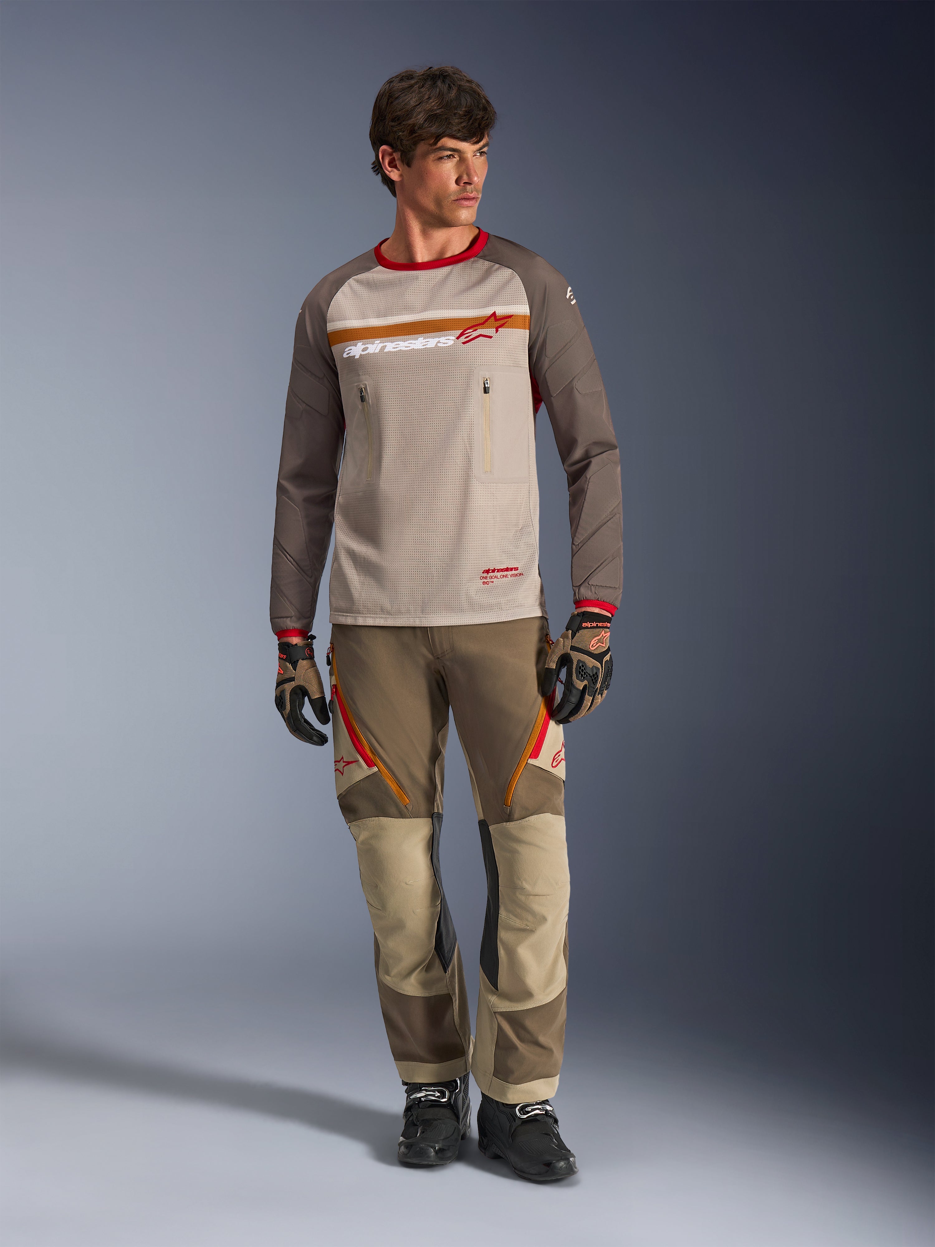 Jersey Alpinestars Maxdura Dual en Aluminum Walnut, jersey de motociclismo de aventura con torso de malla beige y mangas marrones, con cuello rojo y franja horizontal en el pecho con el logo de Alpinestars, combinado con pantalones de aventura y guantes a juego.