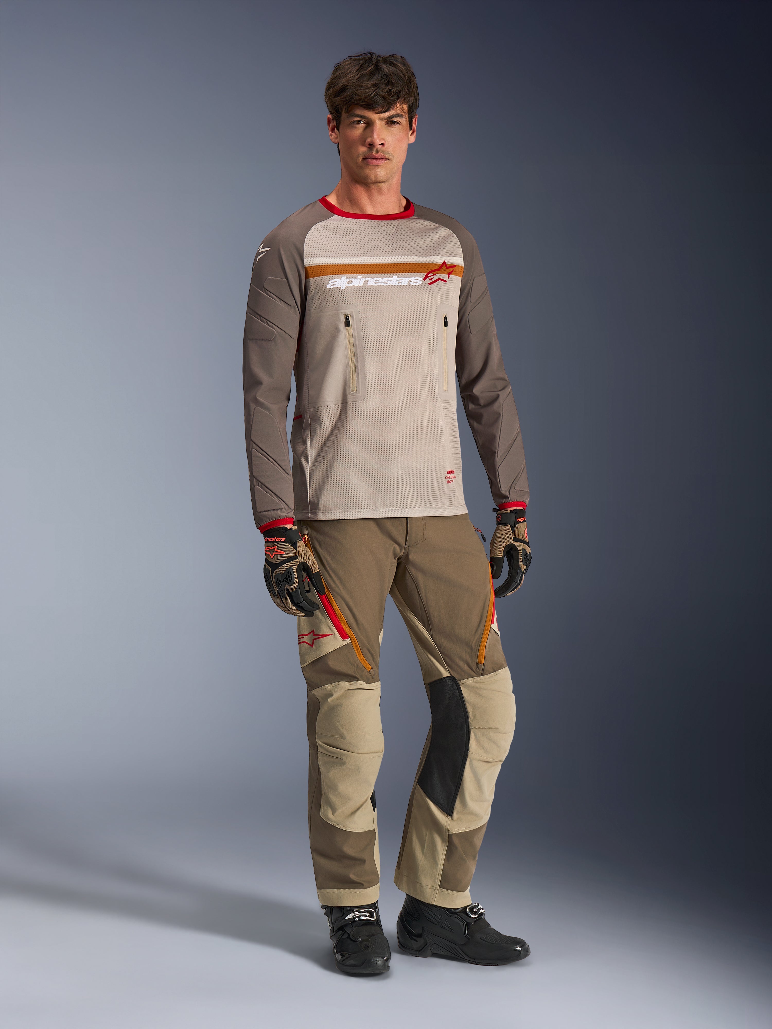 Modelo luciendo el Alpinestars Maxdura Dual Jersey en color Aluminum Walnut, con torso de malla gris, mangas marrones con puños rojos y bolsillos con cremallera en el pecho, combinado con equipamiento off-road a juego para conducción de aventura.