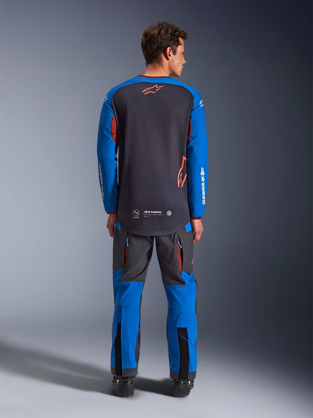 Alpinestars Maxdura Dual Jersey en gris oscuro y azul real, vista trasera que muestra el torso de malla completa y el logotipo de la estrella naranja, mangas azul real con marca blanca, usado con pantalones de aventura grises y azules a juego, equipamiento de alto rendimiento para conducción dual-sport