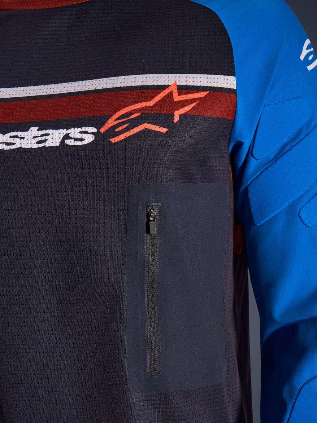 Alpinestars Maxdura Dual Jersey, primer plano de pecho y manga, gris oscuro y azul real, con bolsillo con cremallera en el pecho, torso de malla transpirable y acolchado integrado en las mangas, con el logotipo de Alpinestars sobre franjas rojas y blancas.