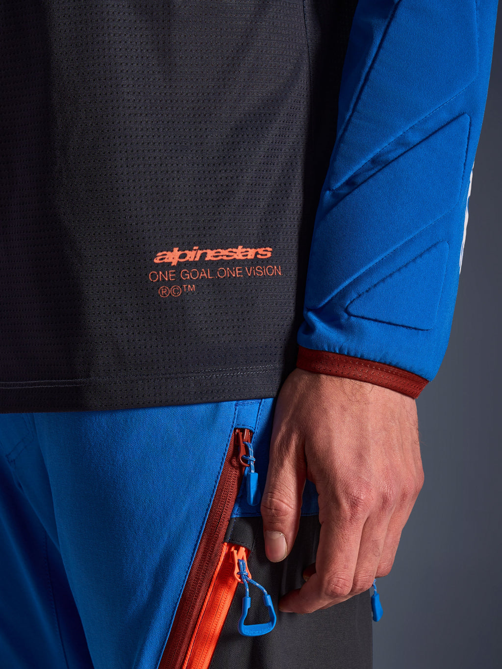 Detalle del jersey Alpinestars Maxdura Dual en gris oscuro y azul real, con logotipo naranja y texto 'One Goal One Vision' en el torso de malla, manga acolchada con paneles geométricos reforzados, equipamiento técnico para conducción de aventura