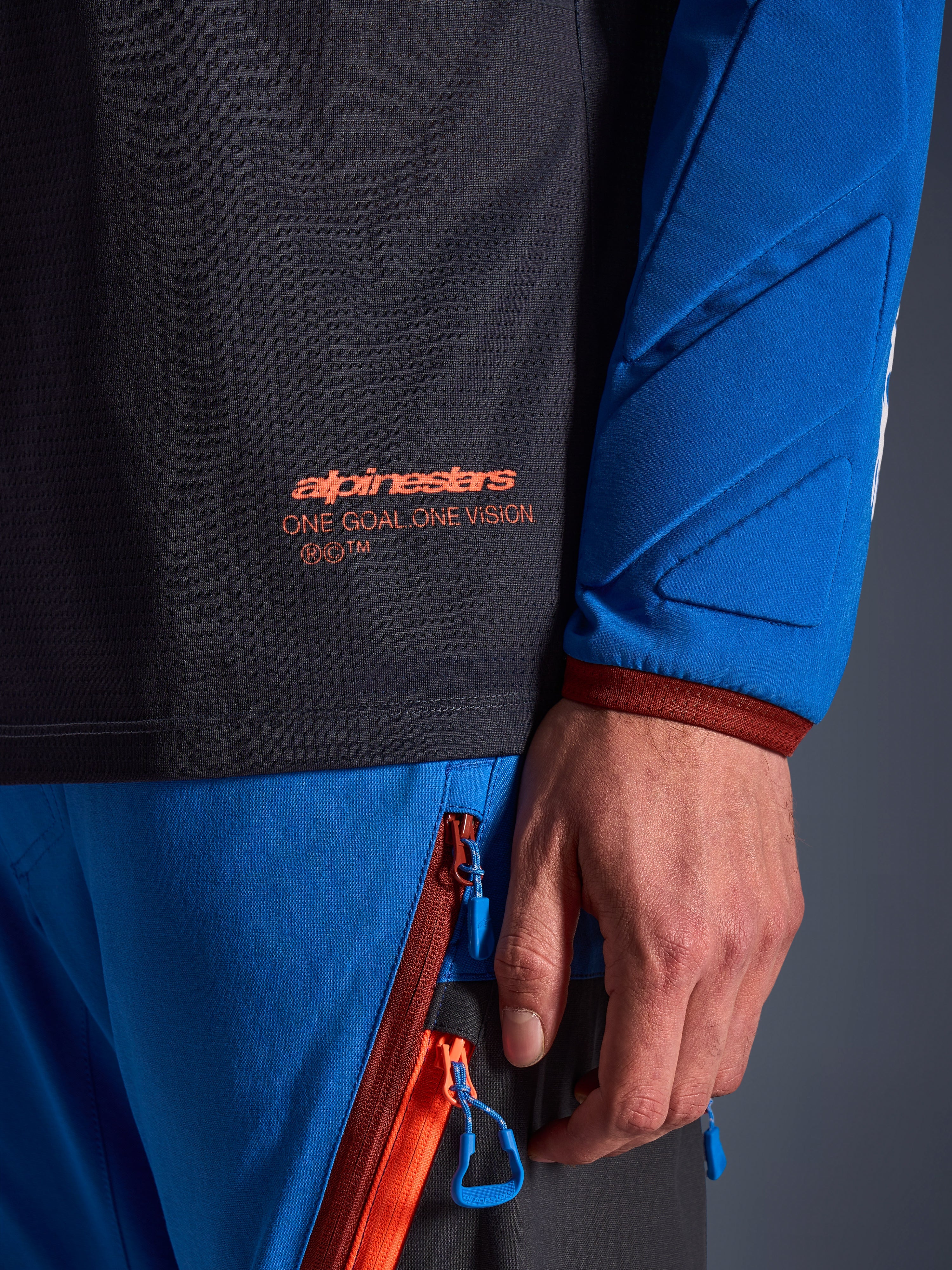 Detalle del jersey Alpinestars Maxdura Dual en gris oscuro y azul real, con logotipo naranja y texto 'One Goal One Vision' en el torso de malla, manga acolchada con paneles geométricos reforzados, equipamiento técnico para conducción de aventura