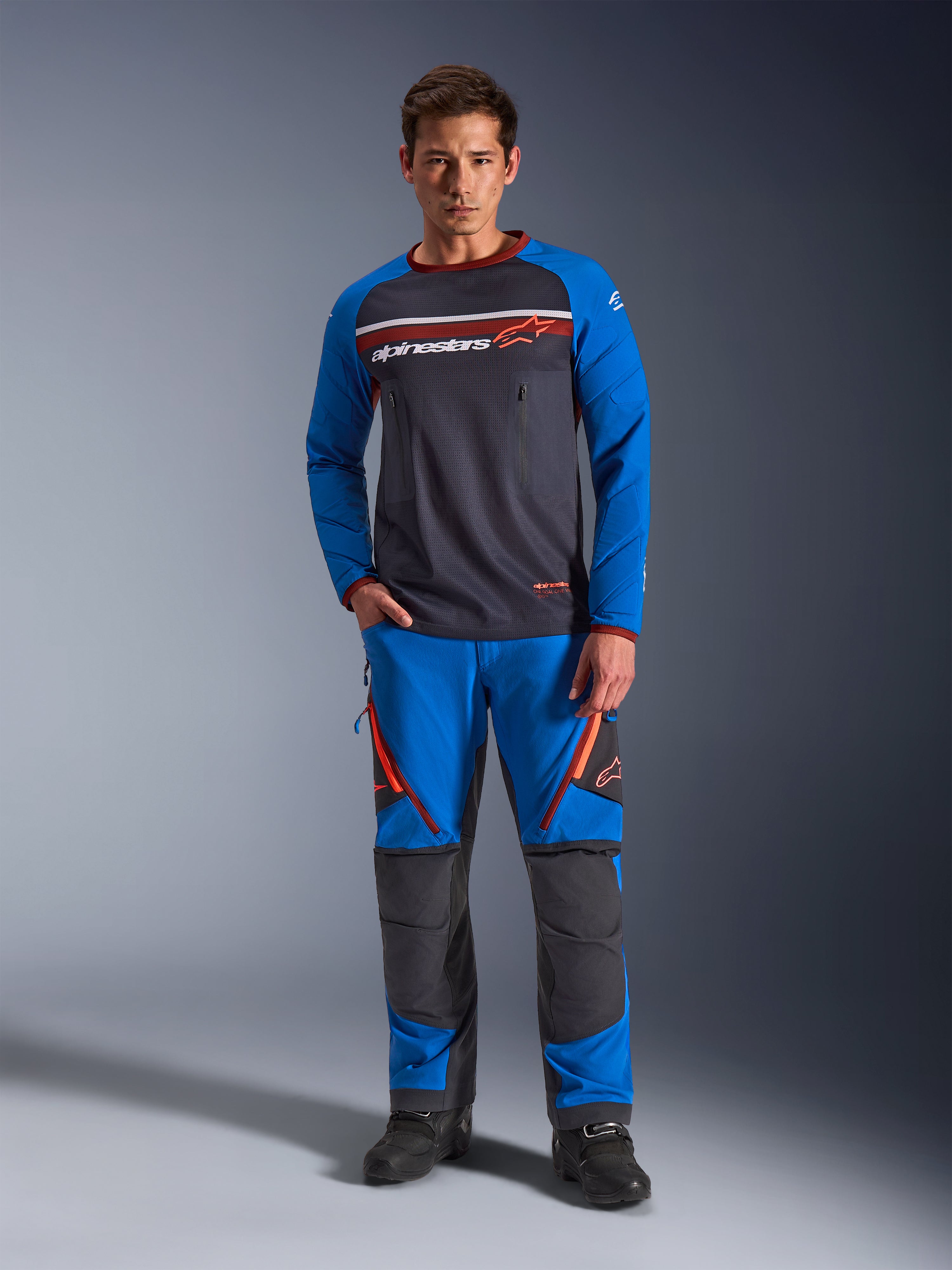 Alpinestars Maxdura Dual Jersey, jersey de motociclismo, gris oscuro y azul real, torso de malla completa con mangas acolchadas y bolsillos con cremallera en el pecho, con logotipo de Alpinestars en blanco y naranja, usado con pantalones de aventura a juego y botas negras