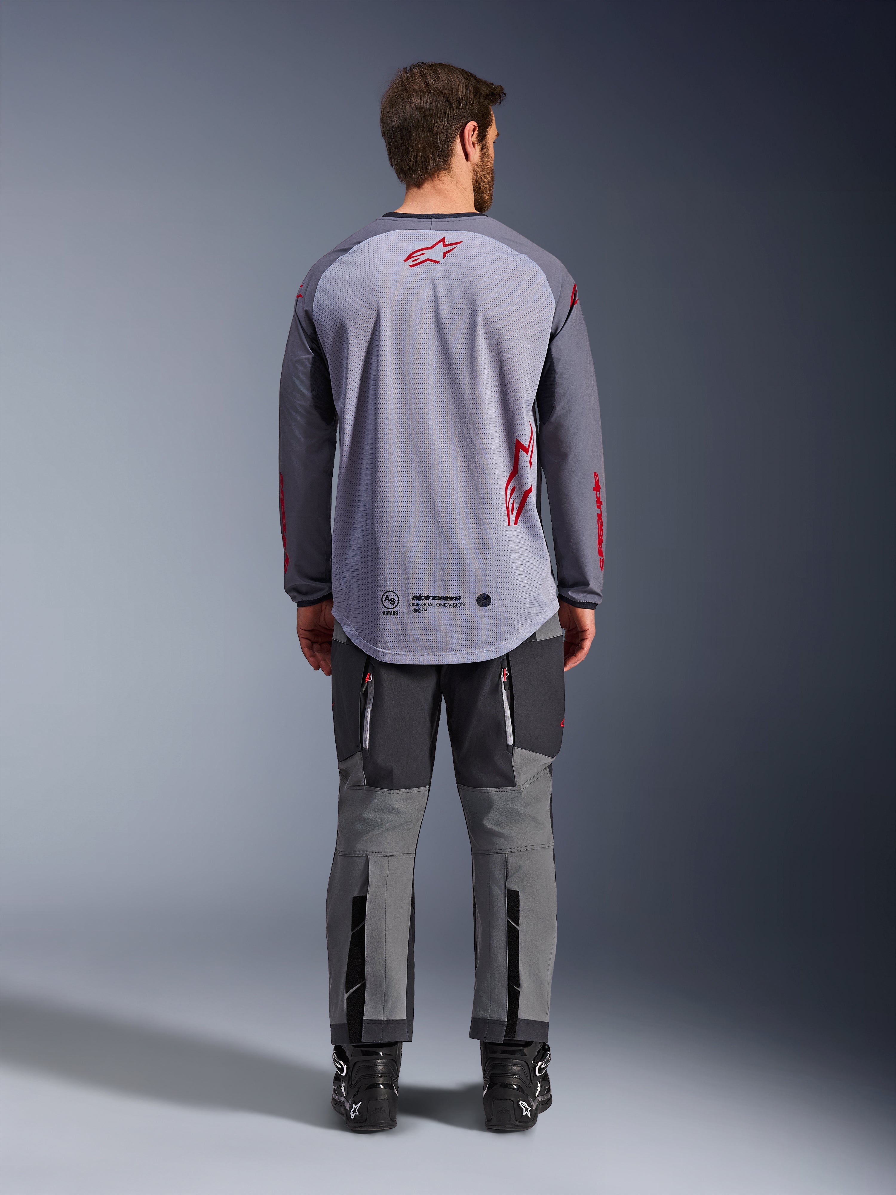 Modelo masculino desde atrás vistiendo la Alpinestars Maxdura Dual Jersey, jersey de aventura en gris medio y gris oscuro con detalles del logo en rojo, construcción de malla en la espalda, combinada con pantalones off-road grises a juego y botas negras