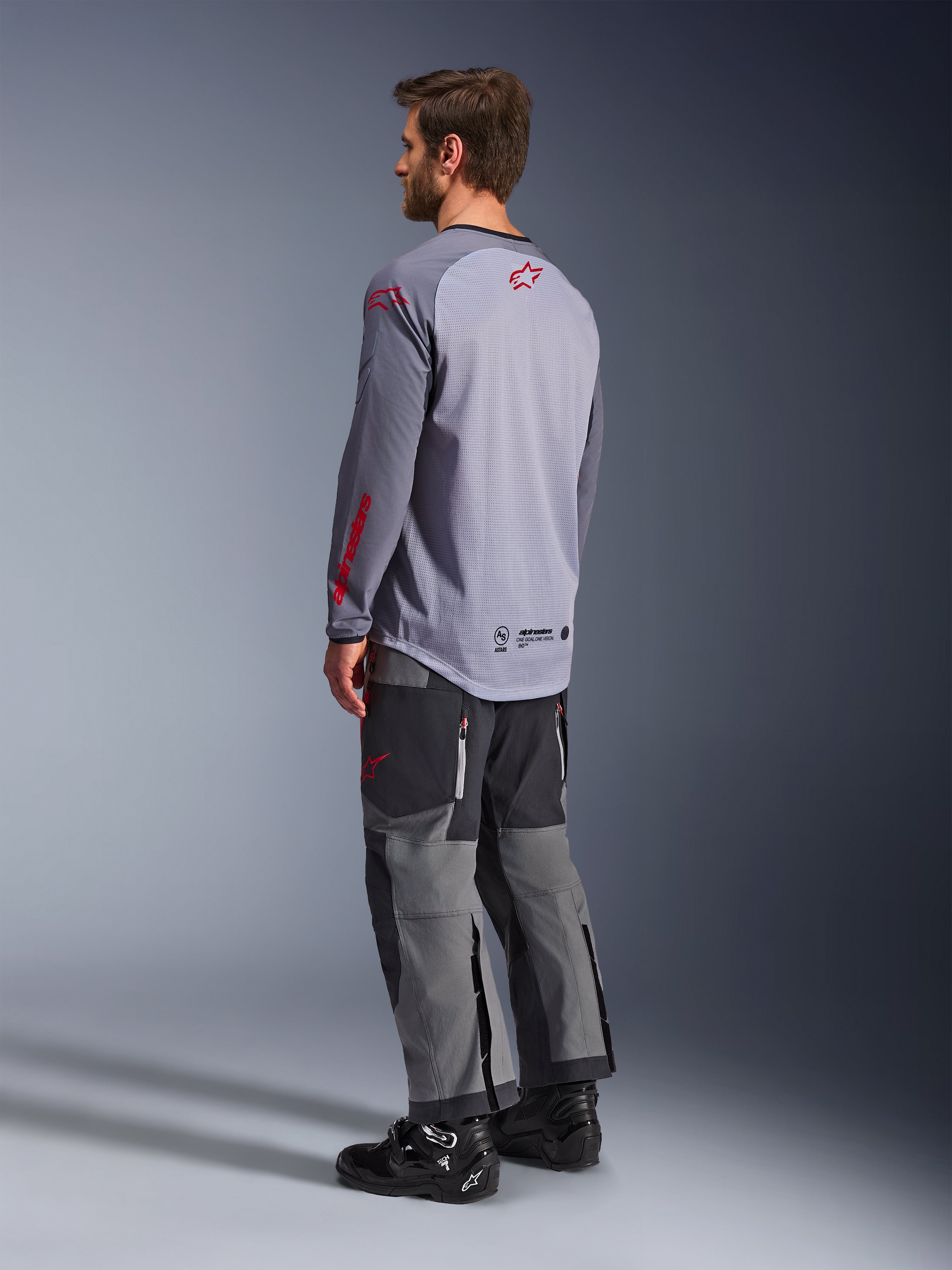 Hombre con el Jersey Alpinestars Maxdura Dual, gris claro con logotipos Alpinestars rojos, vista trasera que muestra el torso de malla y mangas largas, combinado con pantalones técnicos gris carbón y botas de moto negras