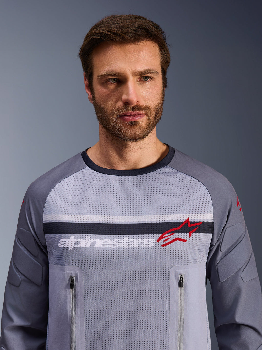 Alpinestars Maxdura Dual Jersey, gris medio y gris oscuro, usada por un modelo masculino, con torso de malla para ventilación, dos bolsillos con cremallera en el pecho, marca 'alpinestars' blanca y logotipo de estrella roja, equipamiento de alto rendimiento para aventura off-road