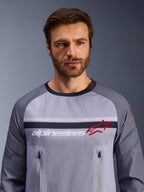 Alpinestars Maxdura Dual Jersey, gris medio y gris oscuro, usada por un modelo masculino, con torso de malla para ventilación, dos bolsillos con cremallera en el pecho, marca 'alpinestars' blanca y logotipo de estrella roja, equipamiento de alto rendimiento para aventura off-road