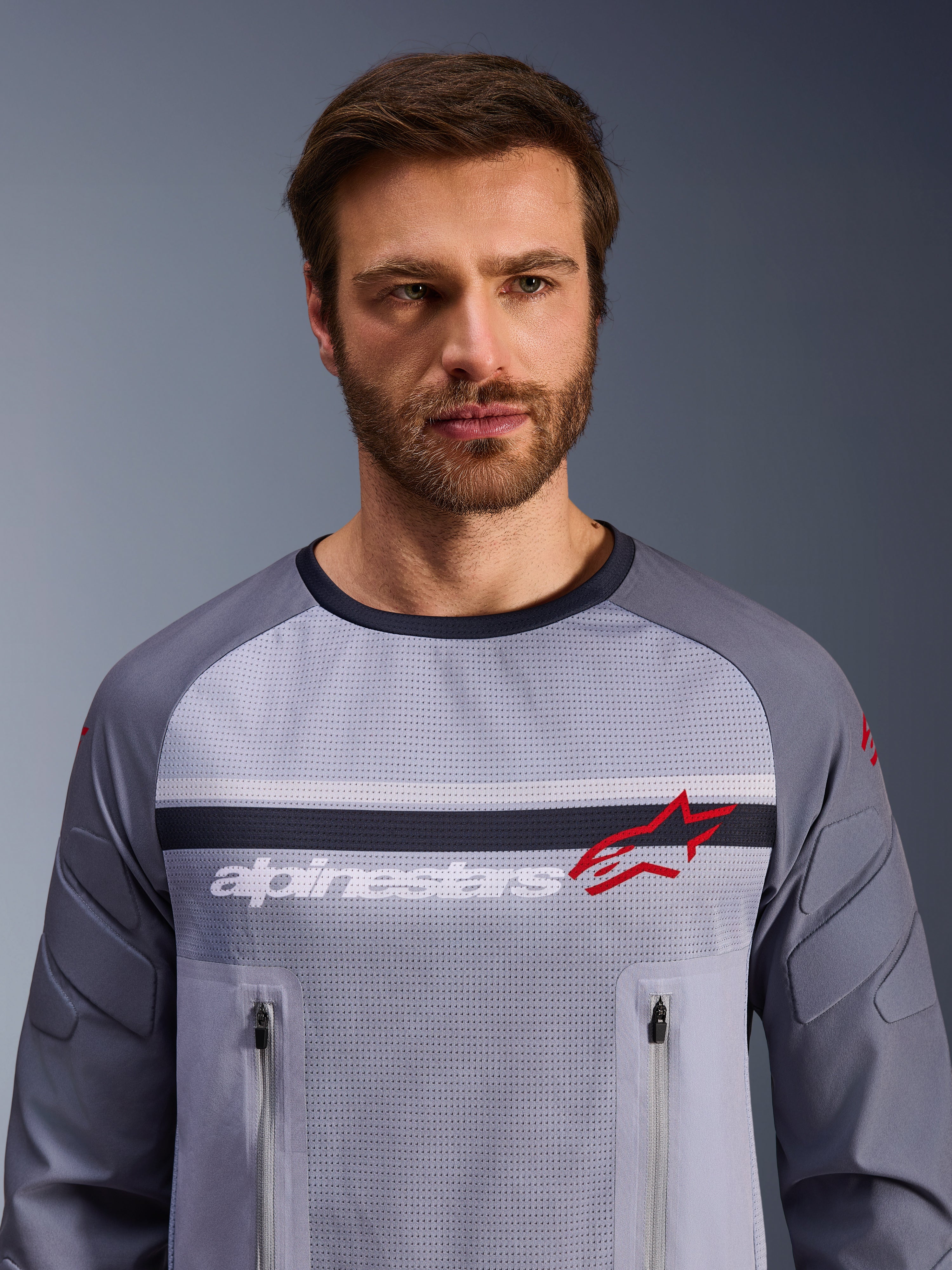 Alpinestars Maxdura Dual Jersey, gris medio y gris oscuro, usada por un modelo masculino, con torso de malla para ventilación, dos bolsillos con cremallera en el pecho, marca 'alpinestars' blanca y logotipo de estrella roja, equipamiento de alto rendimiento para aventura off-road