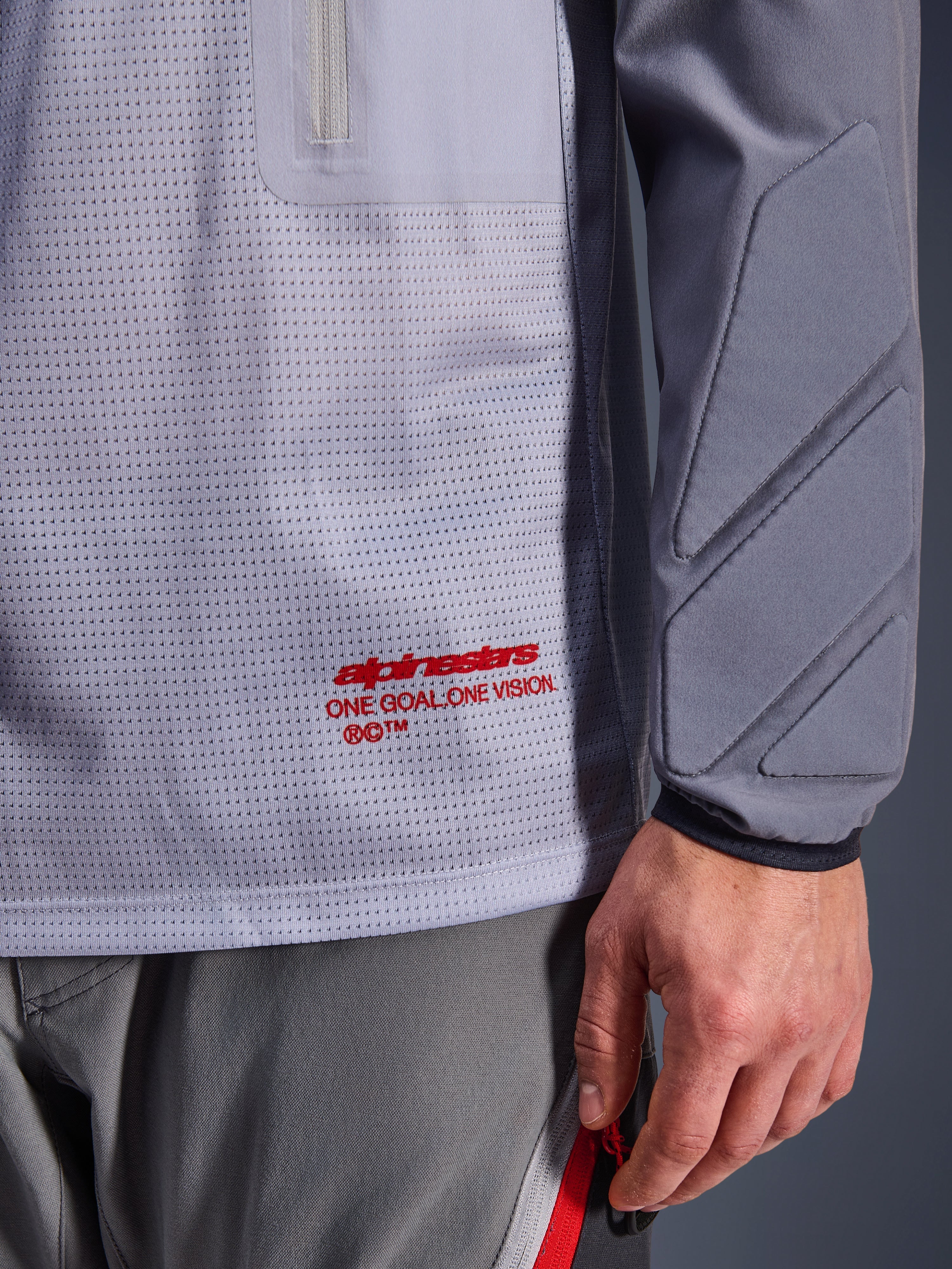Alpinestars Maxdura Dual Jersey, gris medio y gris oscuro, primer plano que muestra el torso de malla transpirable con el logotipo en rojo, detalle de bolsillo con cremallera y acolchado integrado en la manga, equipamiento técnico de motociclismo de aventura
