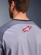 Alpinestars Maxdura Dual Jersey en gris medio y gris oscuro, primer plano de la parte superior de la espalda y el cuello, incluye un pequeño logotipo rojo de la estrella de Alpinestars sobre tejido de malla transpirable gris claro con ribete del cuello en gris oscuro, diseñado para conducción de aventura off-road