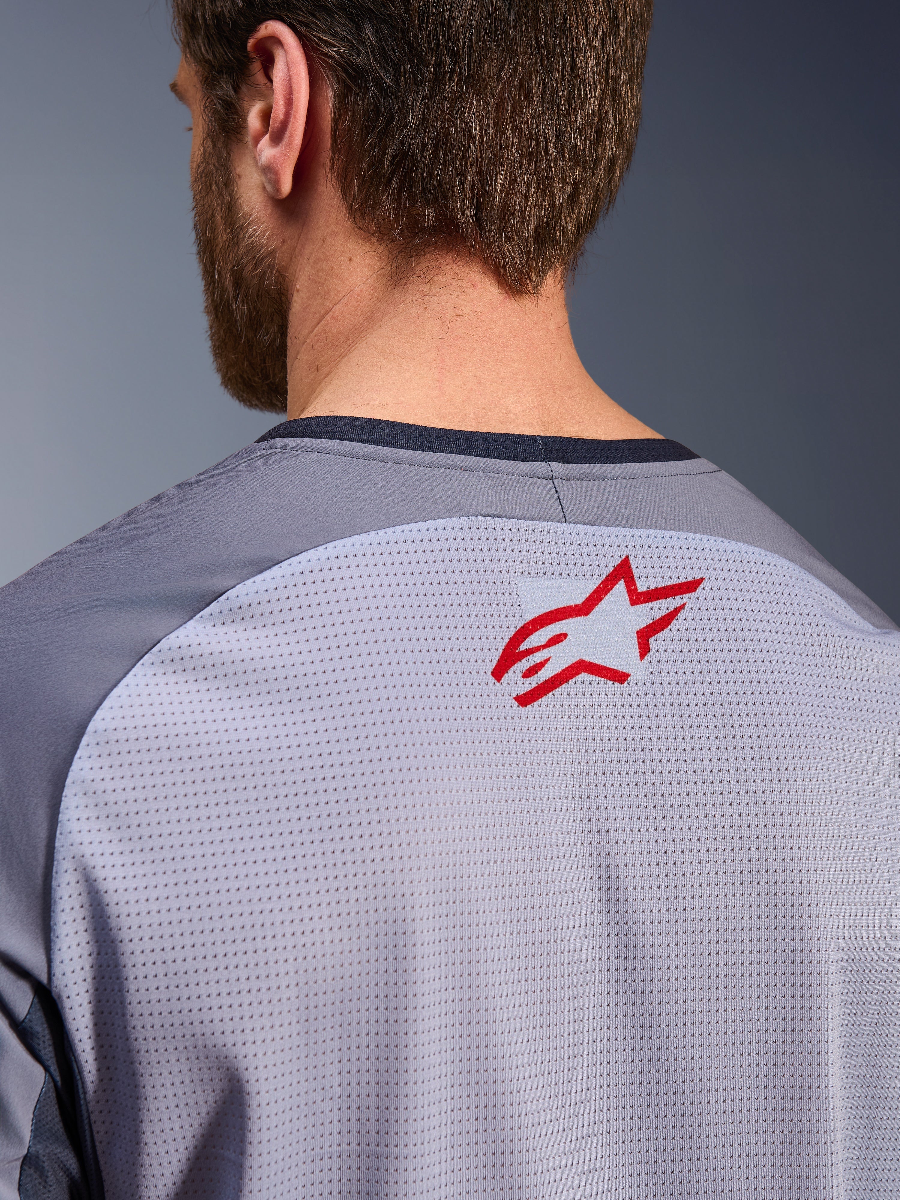 Alpinestars Maxdura Dual Jersey en gris medio y gris oscuro, primer plano de la parte superior de la espalda y el cuello, incluye un pequeño logotipo rojo de la estrella de Alpinestars sobre tejido de malla transpirable gris claro con ribete del cuello en gris oscuro, diseñado para conducción de aventura off-road