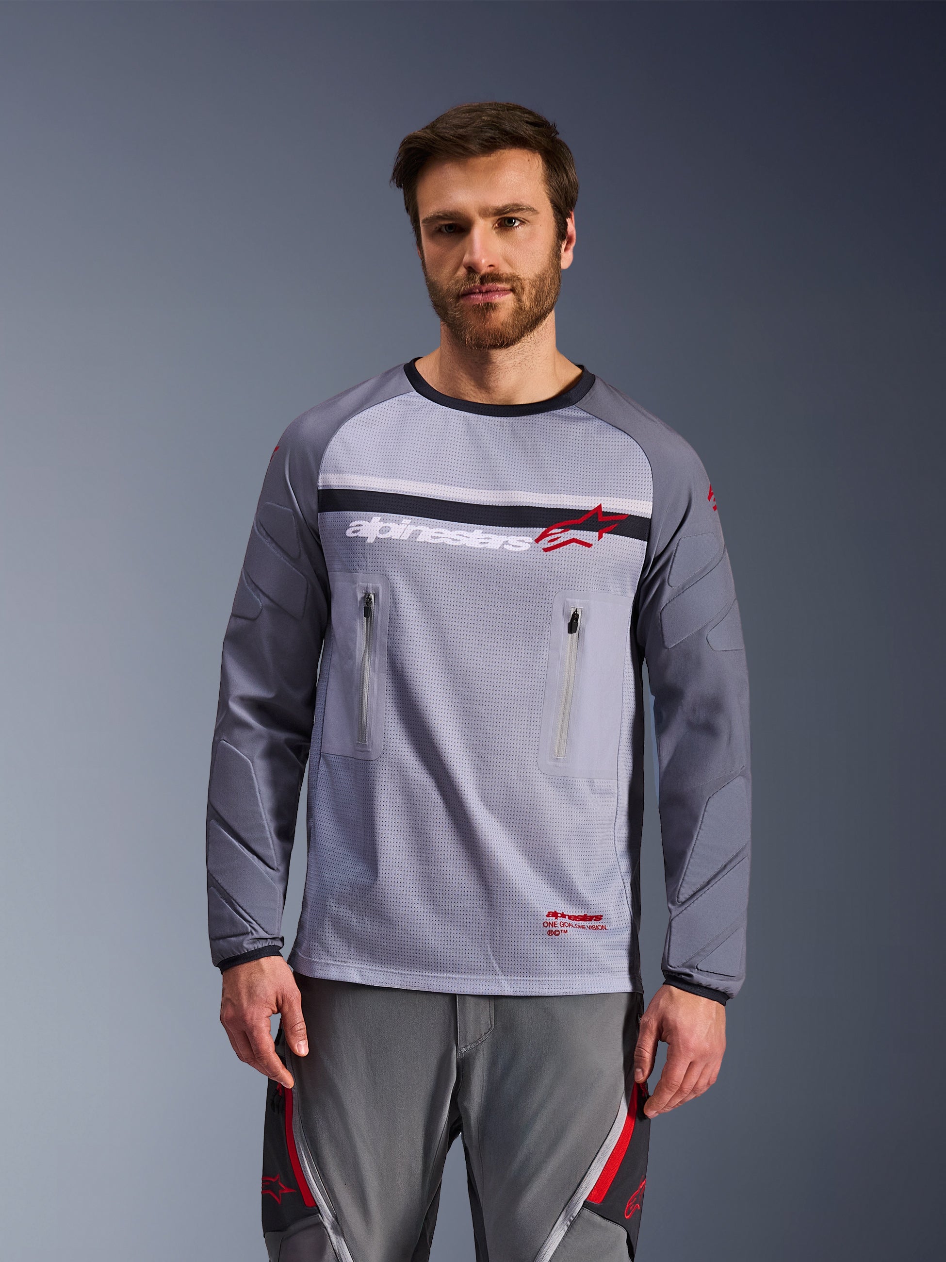 Alpinestars Maxdura Dual Jersey, gris medio y gris oscuro, usado por un modelo con pantalones grises a juego, cuenta con bolsillos con cremallera en el pecho, construcción del torso en malla y mangas acolchadas resistentes a la abrasión, equipo de motociclismo de aventura off-road