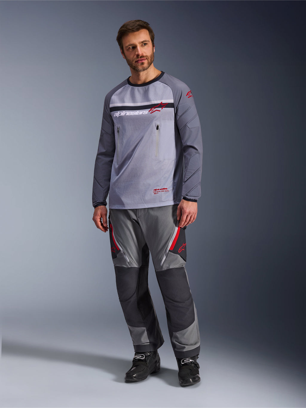Un hombre vistiendo el jersey Alpinestars Maxdura Dual en gris medio y gris oscuro, combinado con pantalones de aventura en gris y negro a juego y botas negras, con un torso de malla transpirable, bolsillos con cremallera en el pecho y detalles del logotipo en rojo.
