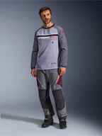 Un hombre vistiendo el jersey Alpinestars Maxdura Dual en gris medio y gris oscuro, combinado con pantalones de aventura en gris y negro a juego y botas negras, con un torso de malla transpirable, bolsillos con cremallera en el pecho y detalles del logotipo en rojo.