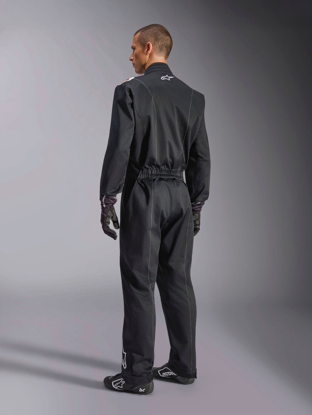Alpinestars Kart Indoor V2 Suit, Black one-piece karting suit shown from a back three-quarter angle, featuring an elastic waist and a white logo on the neck, worn with black racing gloves and shoes, durable design for recreational karting