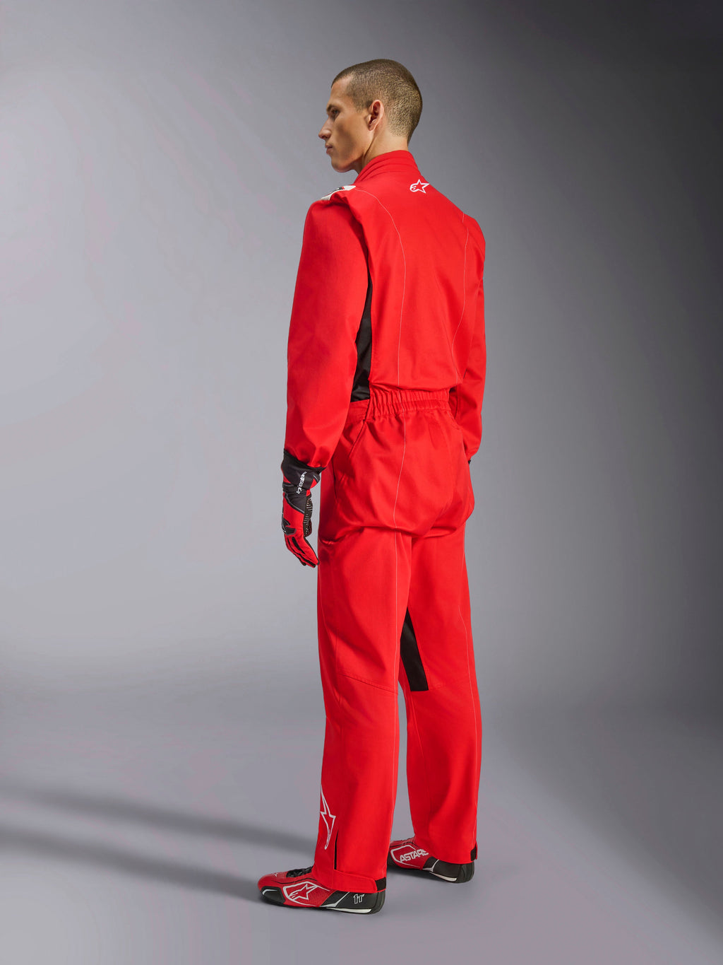 Alpinestars Kart Indoor V2 Suit, mono de karting rojo, vista trasera de tres cuartos en un modelo, usado con guantes rojos y calzado de competición a juego, construcción de sarga de poliéster y algodón de una sola capa, equipamiento profesional para karting en interiores