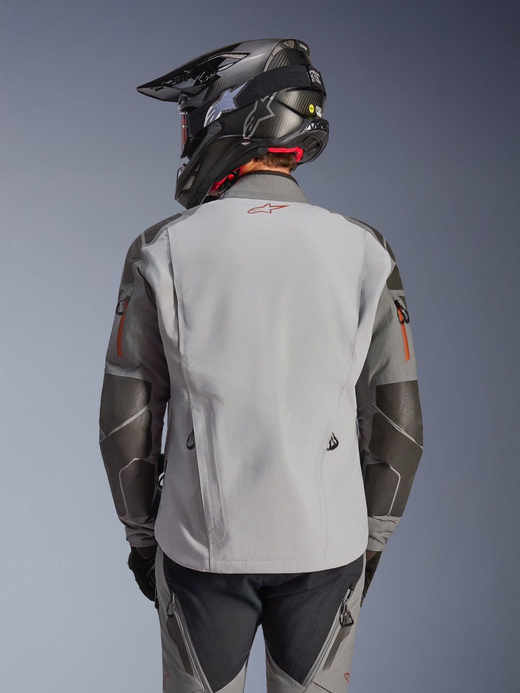 Chaqueta Alpinestars Nazca 3L GORE-TEX PRO, chaqueta de moto de aventura, vista trasera, gris oscuro y gris medio, cuenta con refuerzos Superfabric en codos y hombros, logo rojo en la parte trasera, combinada con equipamiento de aventura a juego y un casco de fibra de carbono.